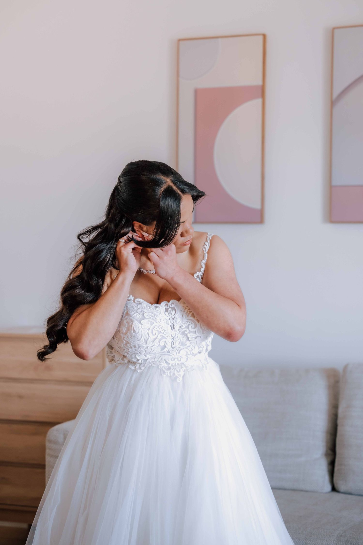 A bride in a white lace ball gown adjusts her earring in a bright modern room.