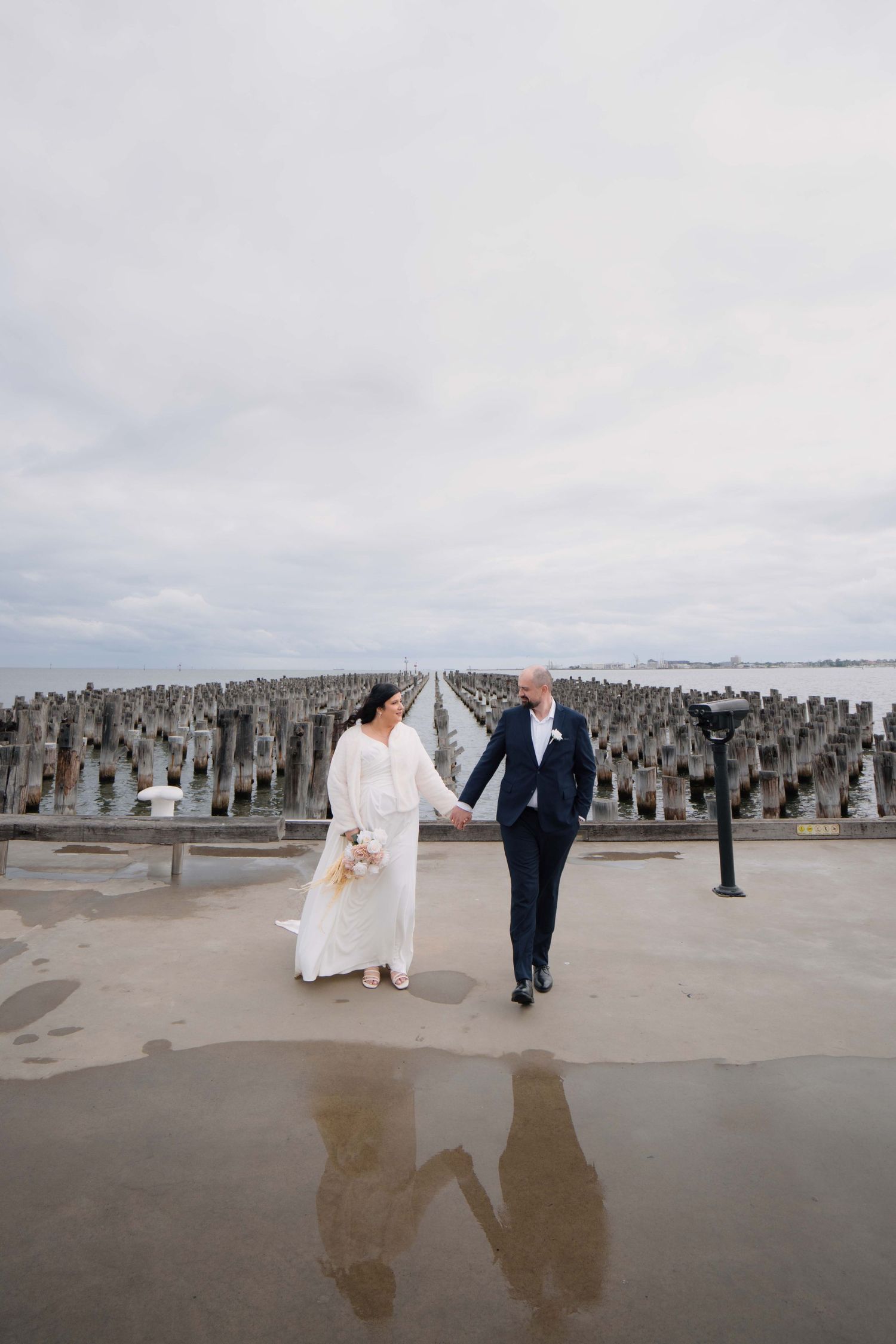 Bride and groom walking hand in hand on a wet pier with overcast skies behind them.