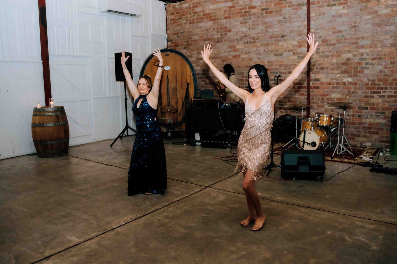 Two women dancing with arms raised in rustic barn venue with brick wall background.