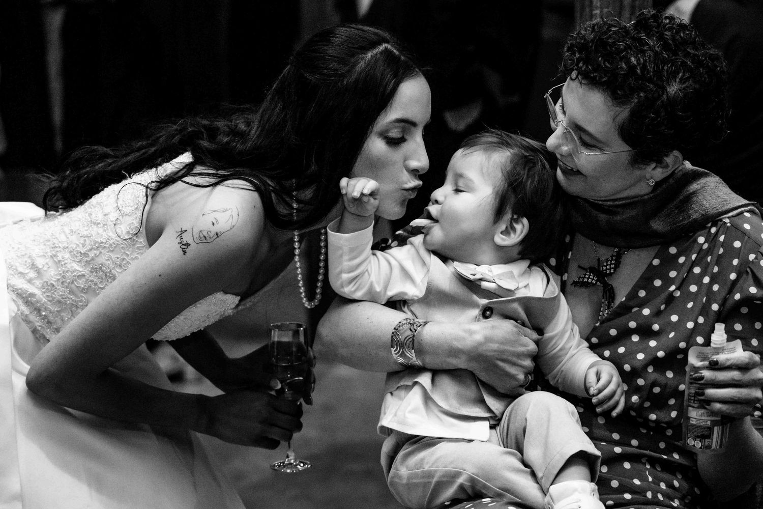 Bride in white lace dress kisses a baby held by a guest in a black and white wedding candid photo.