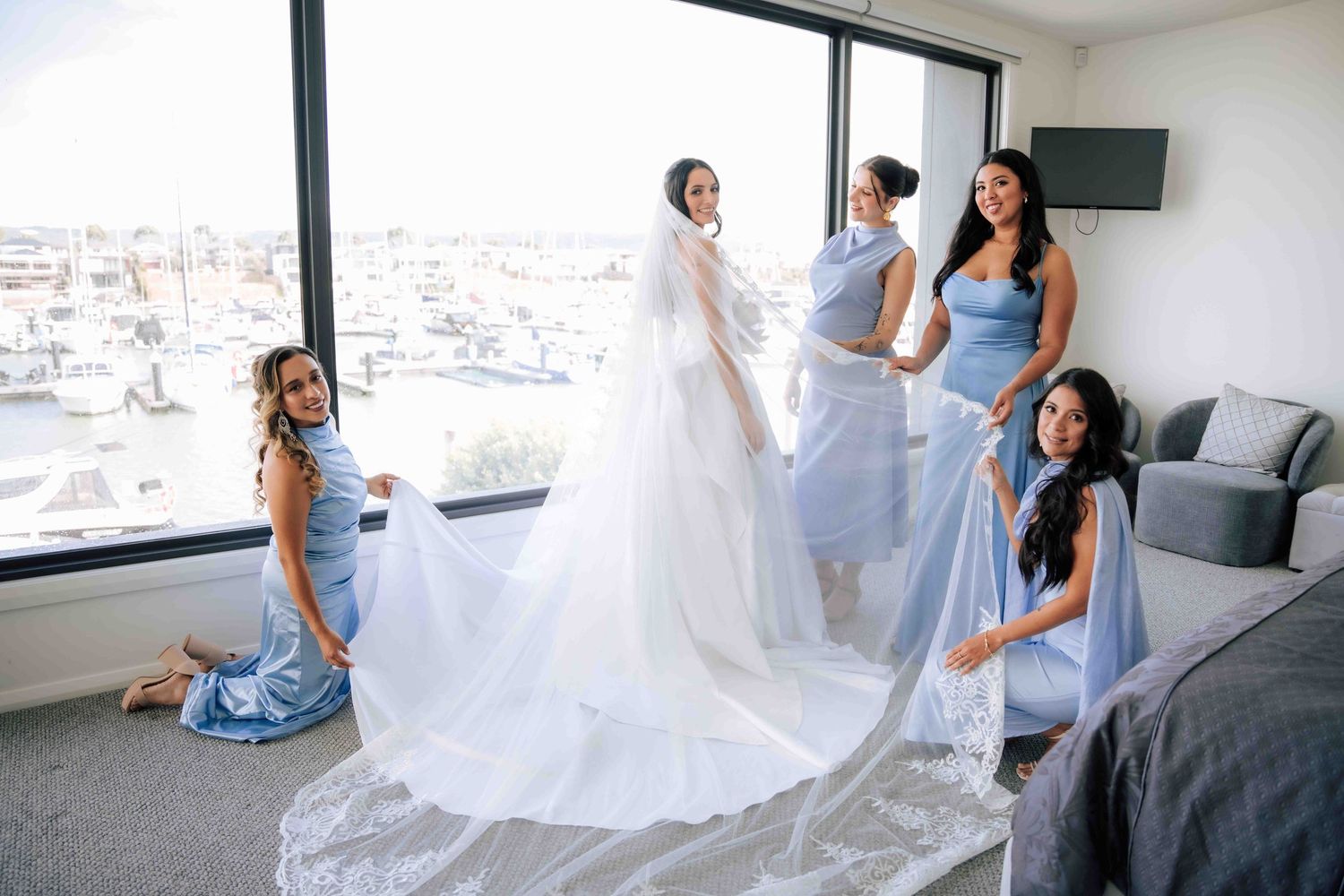 Bride in elegant white gown with long veil surrounded by bridesmaids in light blue dresses in a hotel room.