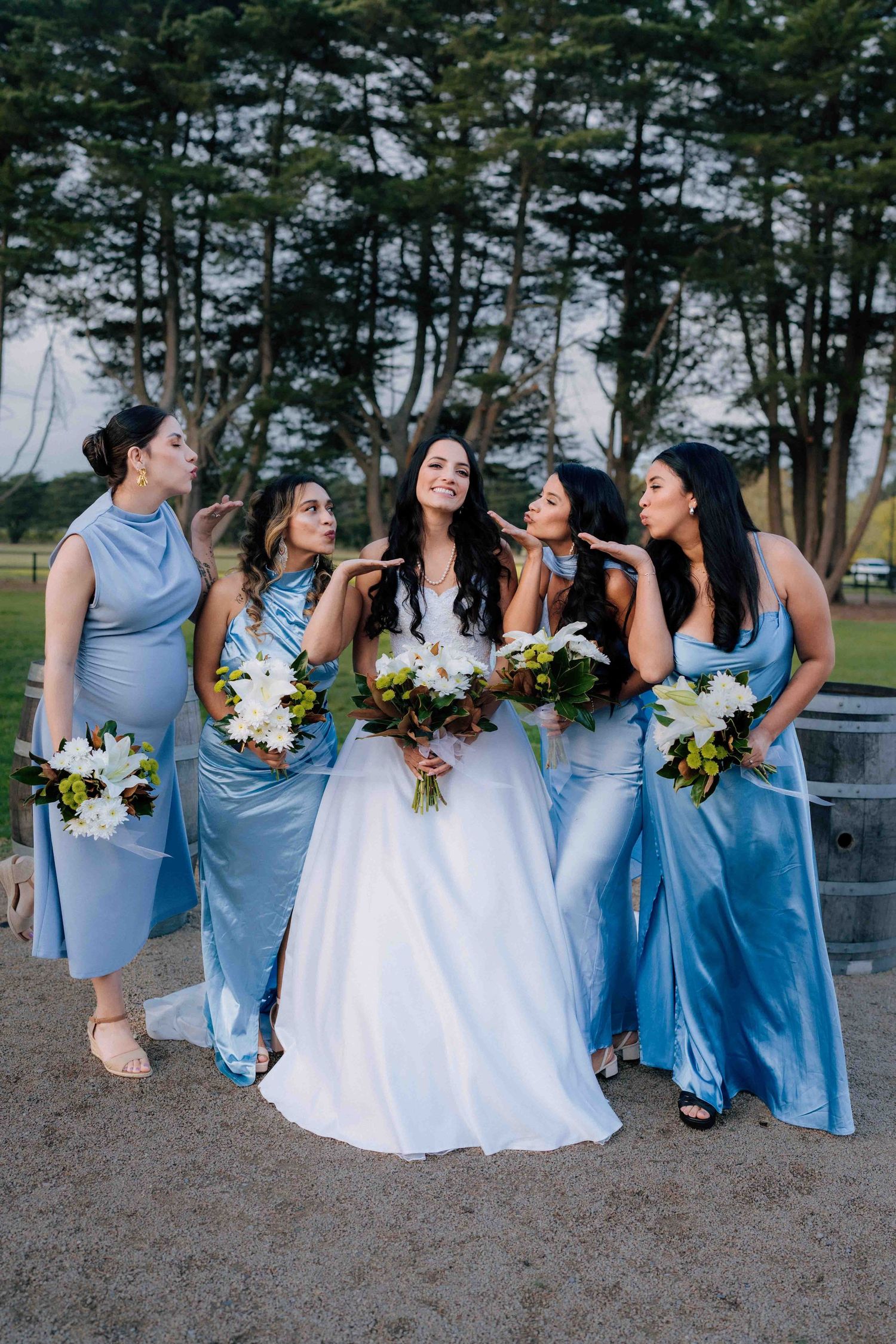 Bride in white gown with bridesmaids in blue satin dresses holding bouquets outdoors.