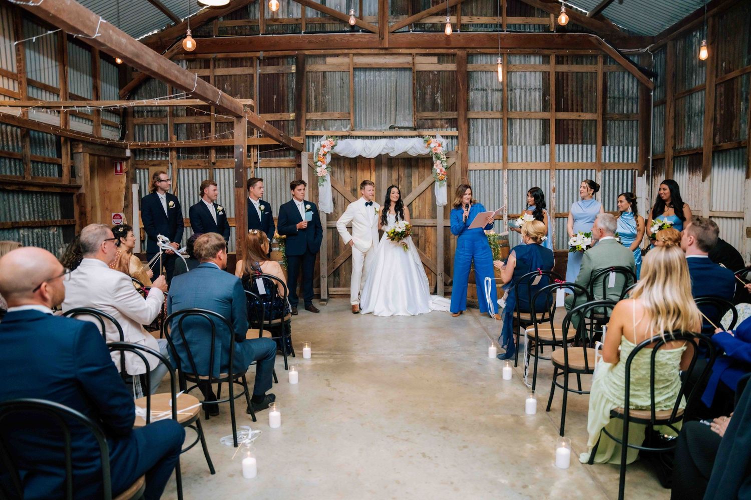 Rustic barn wedding ceremony with bride and groom at floral arch, guests seated in black chairs on concrete floor.