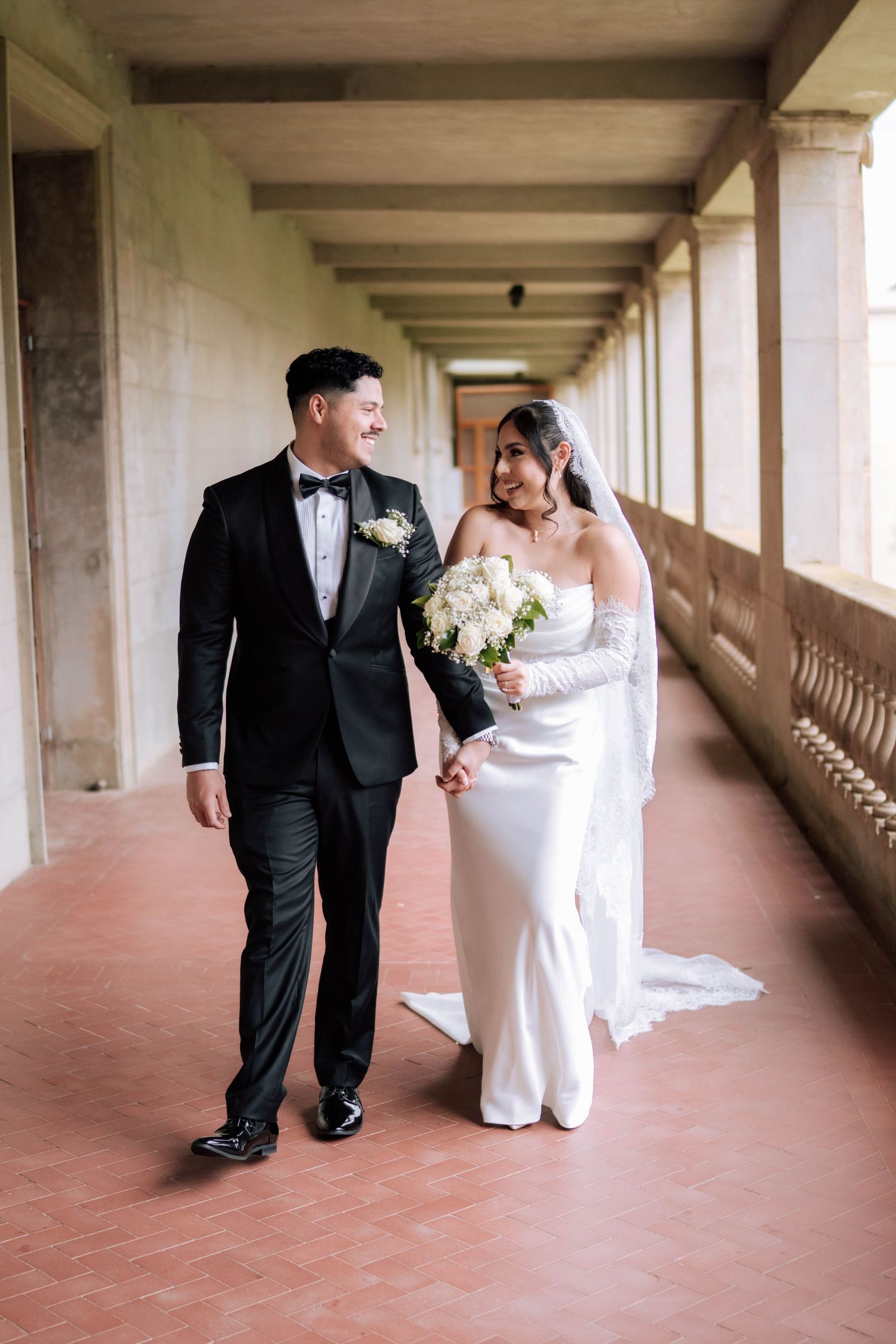 Bride and groom walking hand in hand through elegant columned corridor on wedding day.