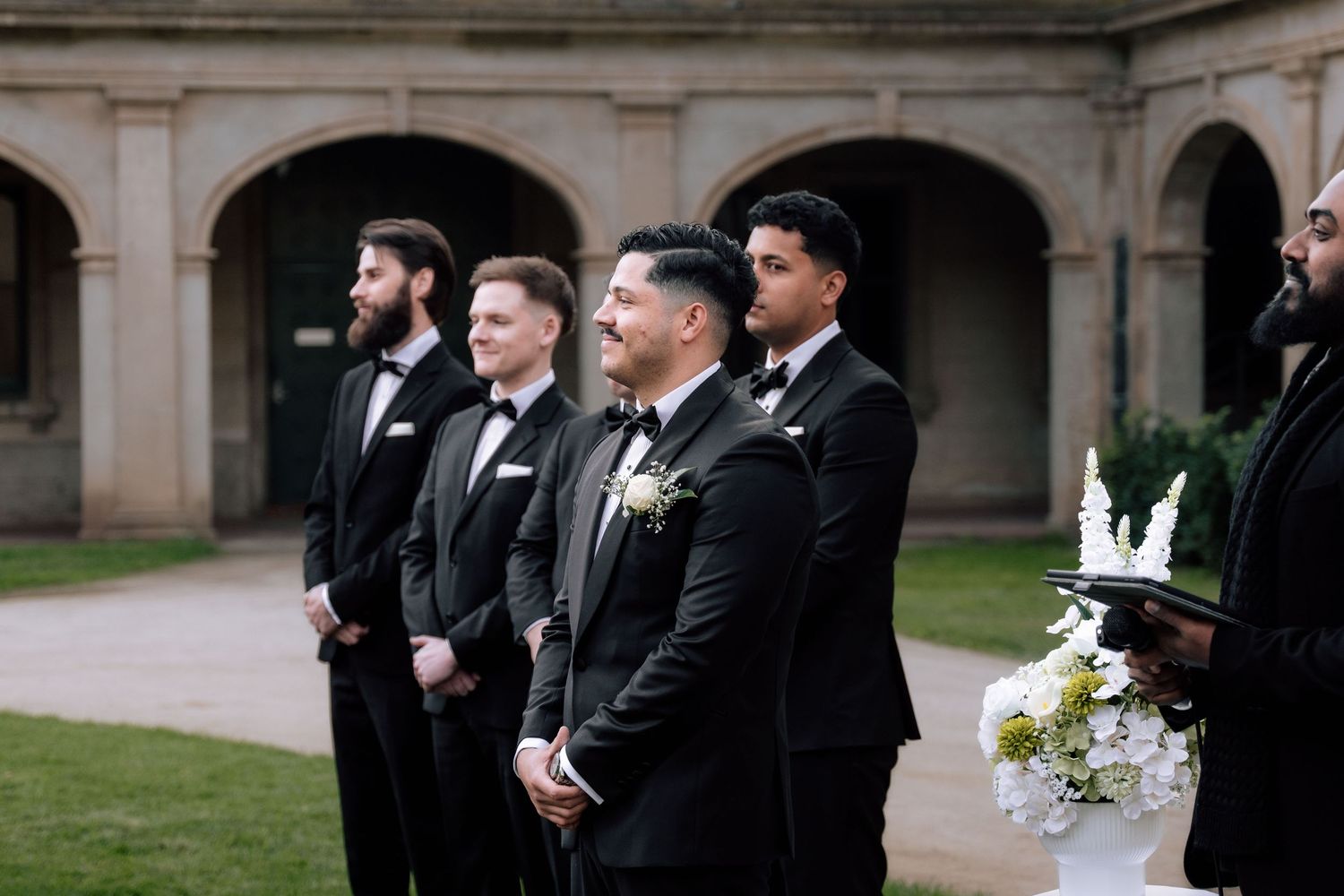 Groom and groomsmen in black tuxedos standing at outdoor wedding ceremony.