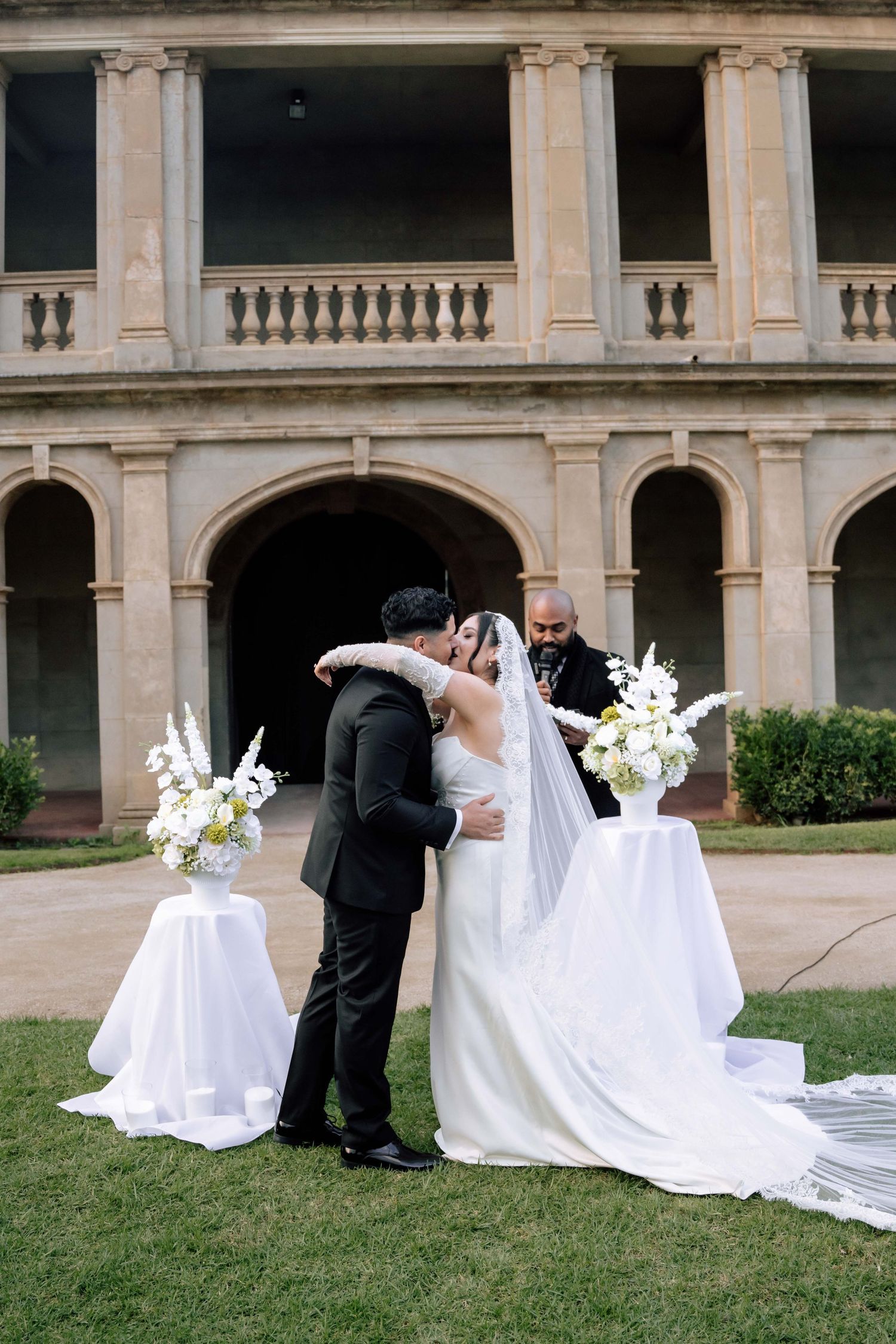 Bride and groom share first kiss at outdoor wedding ceremony before grand historic stone building.
