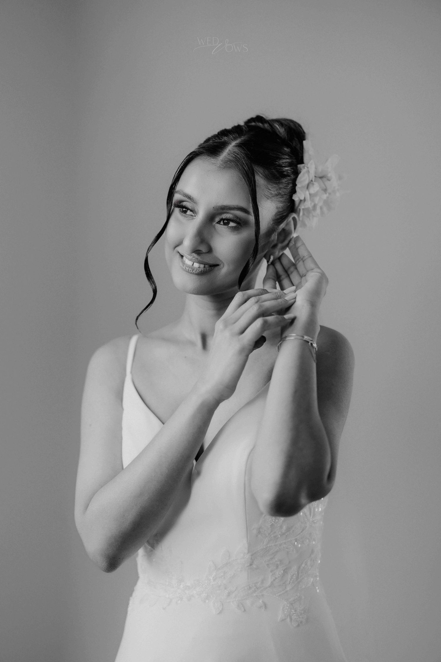 Black and white portrait of a smiling bride in white gown adjusting her floral hair accessory.