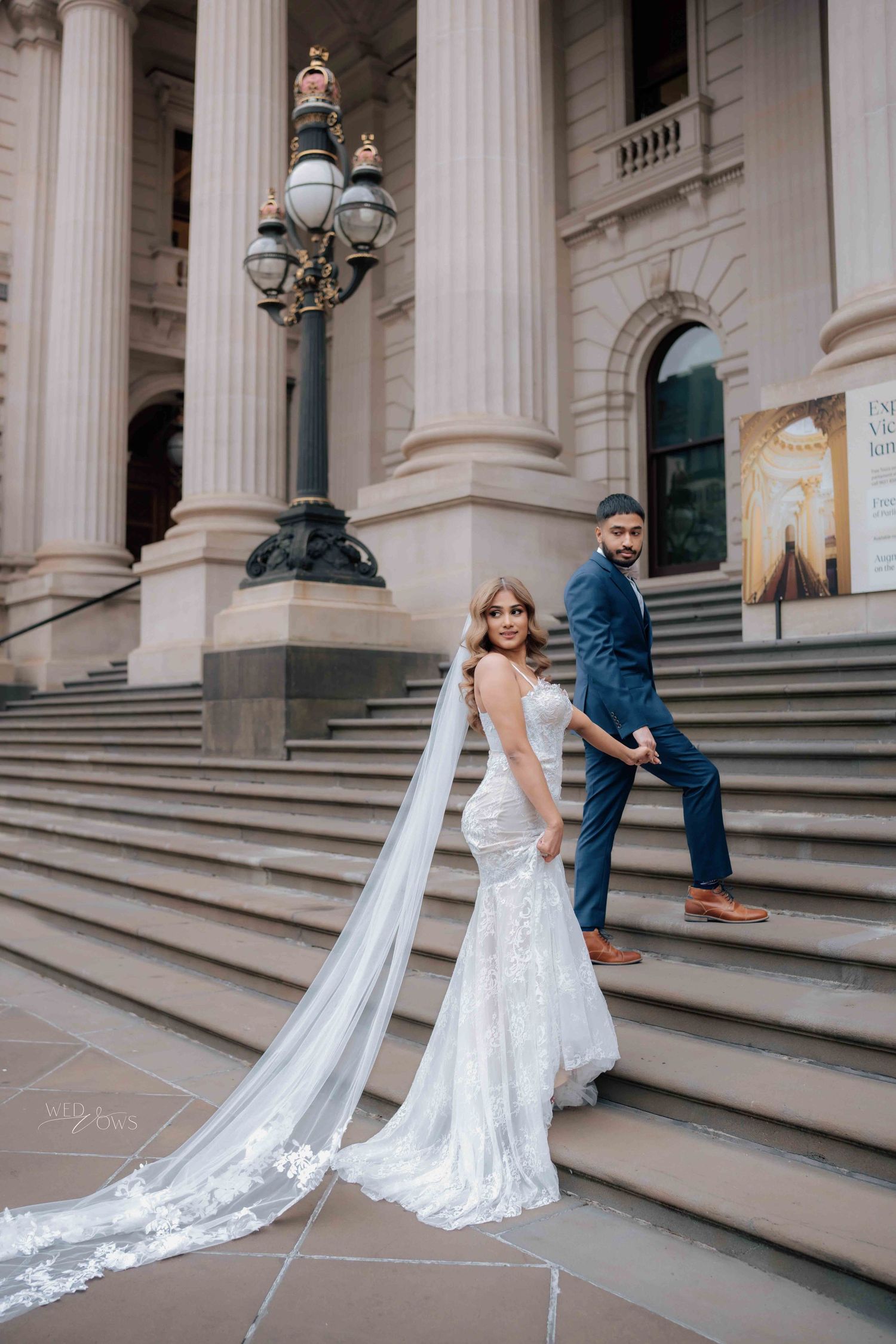 Bride and groom pose on steps of grand building with long cathedral veil trailing.