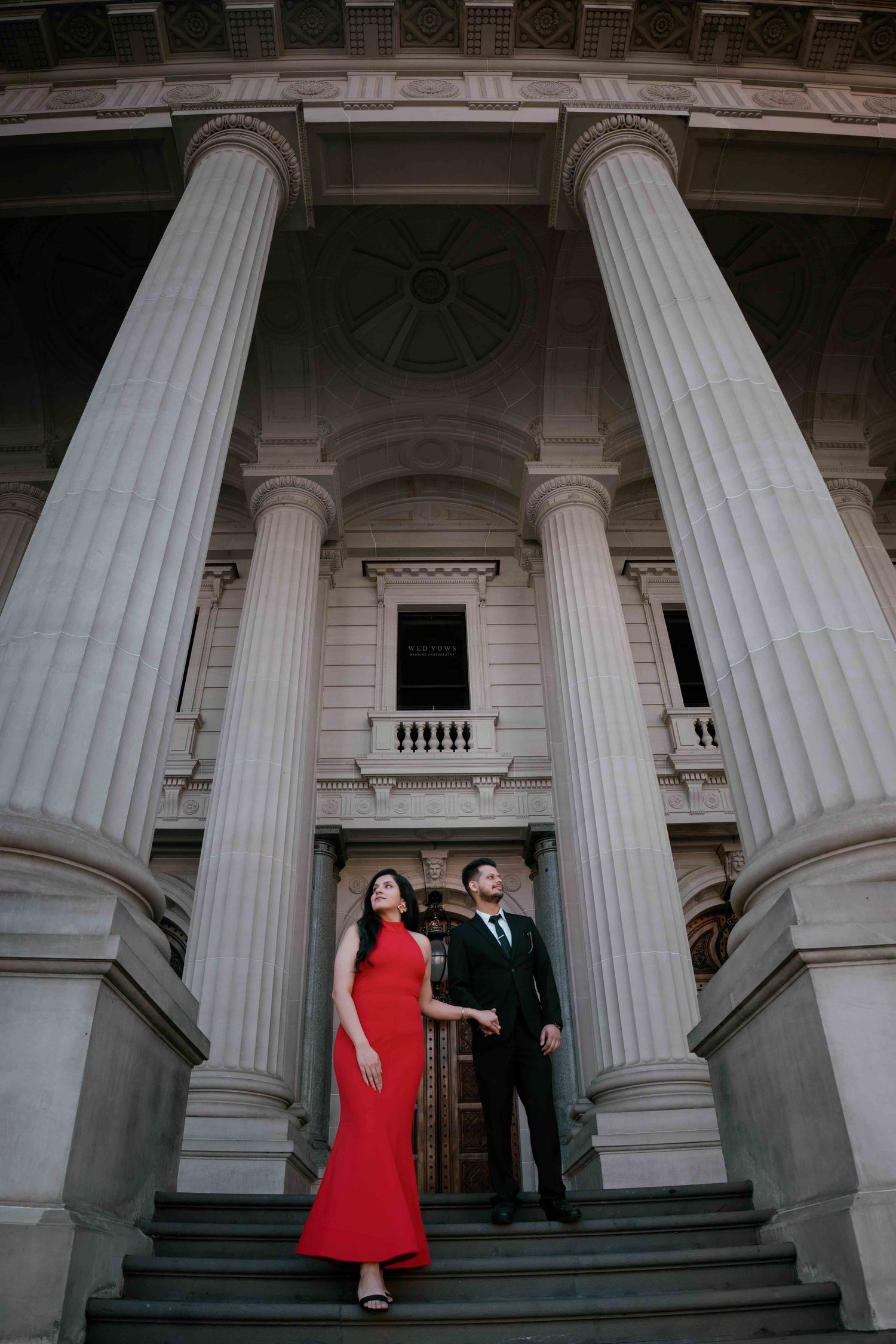 Couple holding hands on steps of majestic columned building, woman in striking red mermaid gown.