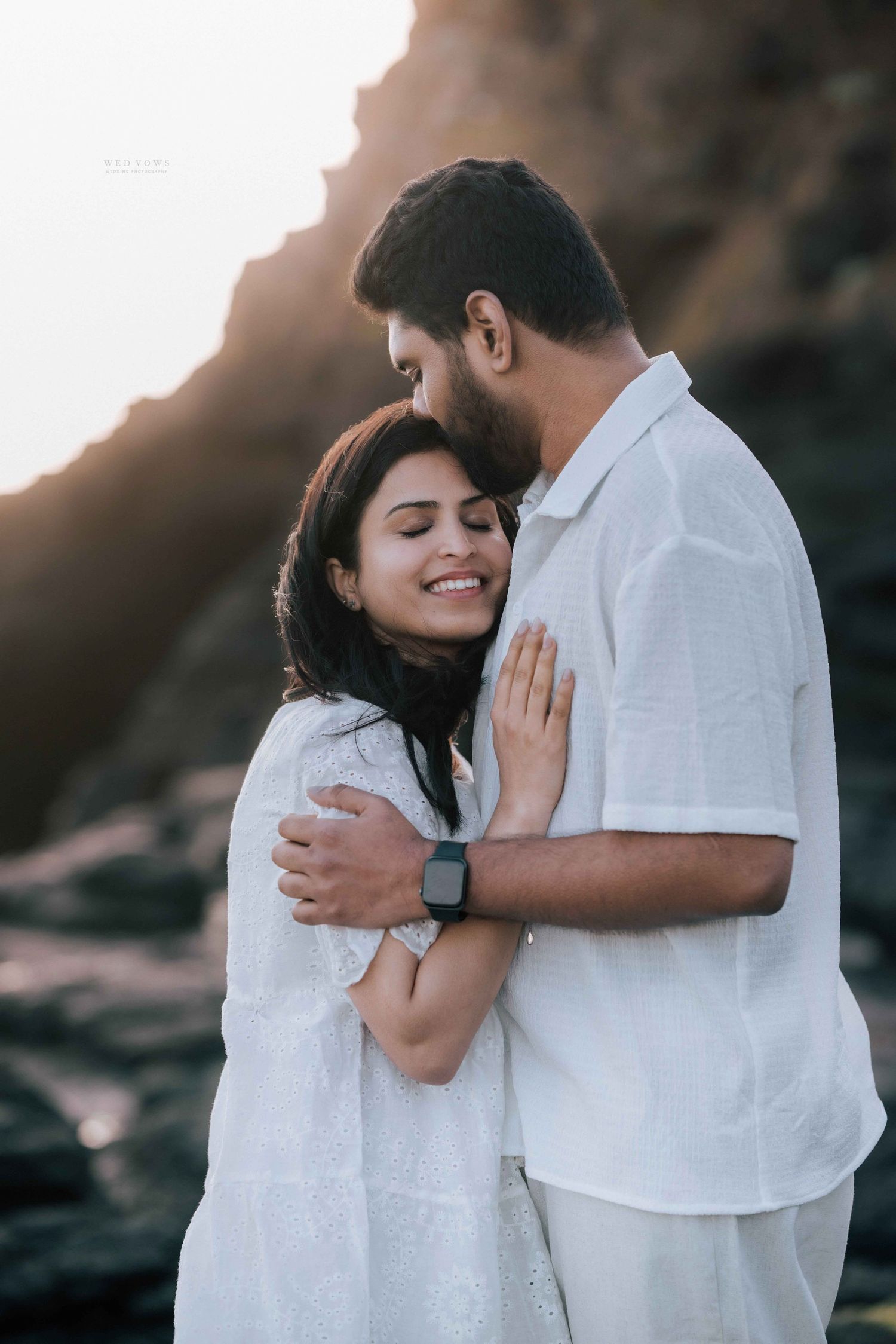 Close-up of smiling couple in white sharing a tender embrace against rocky coastal backdrop.