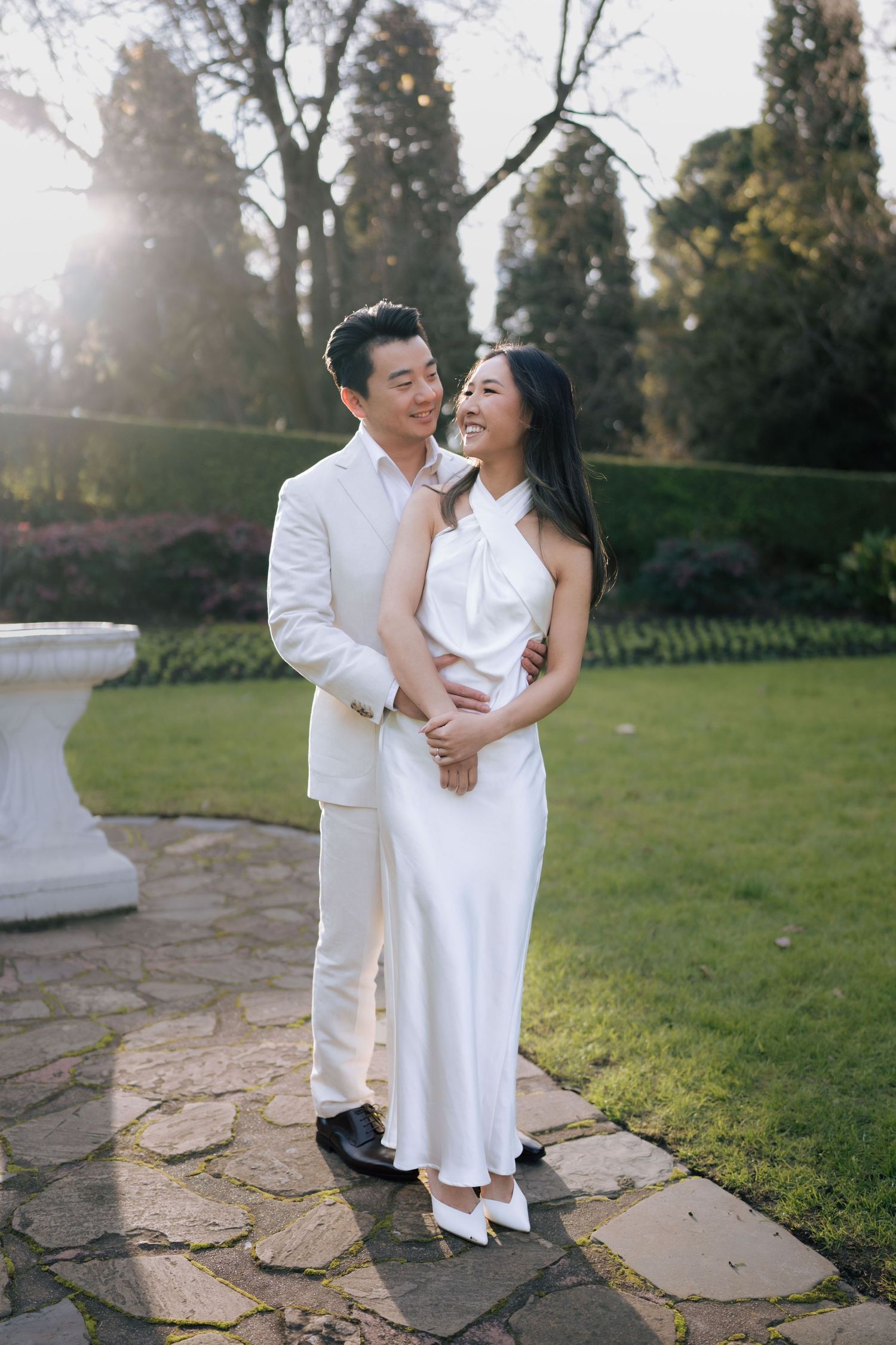 Smiling couple in elegant white outfits embracing on a stone path in a lush garden during golden hour.