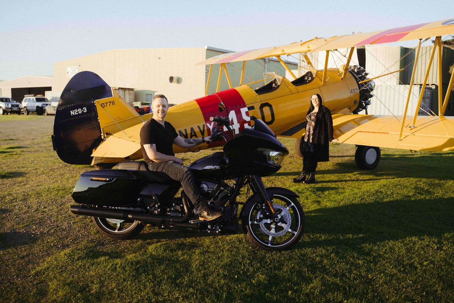 A Harley-Davidson motorcycle parked in front of a vintage yellow biplane on a green airfield with two people nearby.