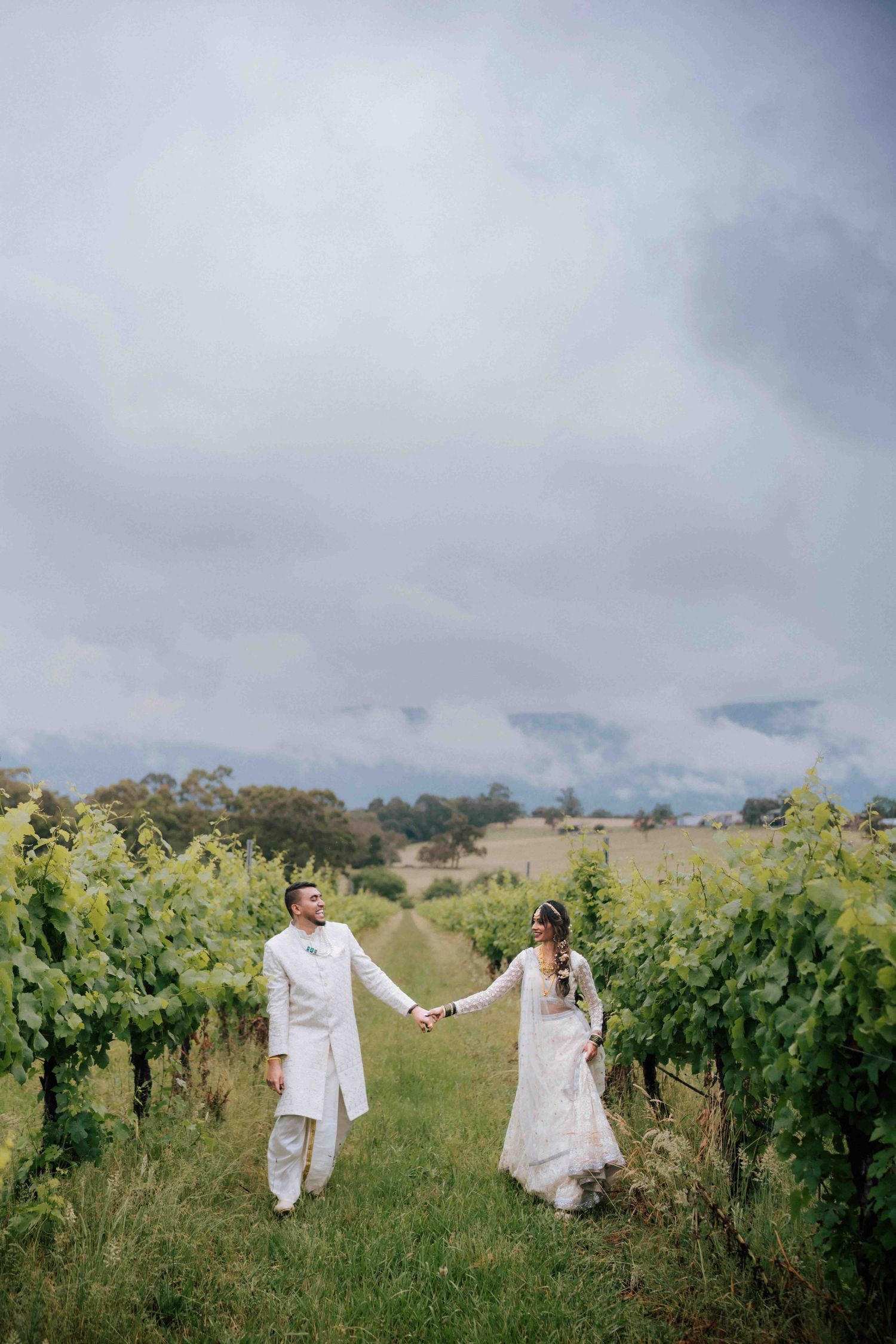 Bride and groom in white attire holding hands while walking through a lush vineyard under cloudy skies.