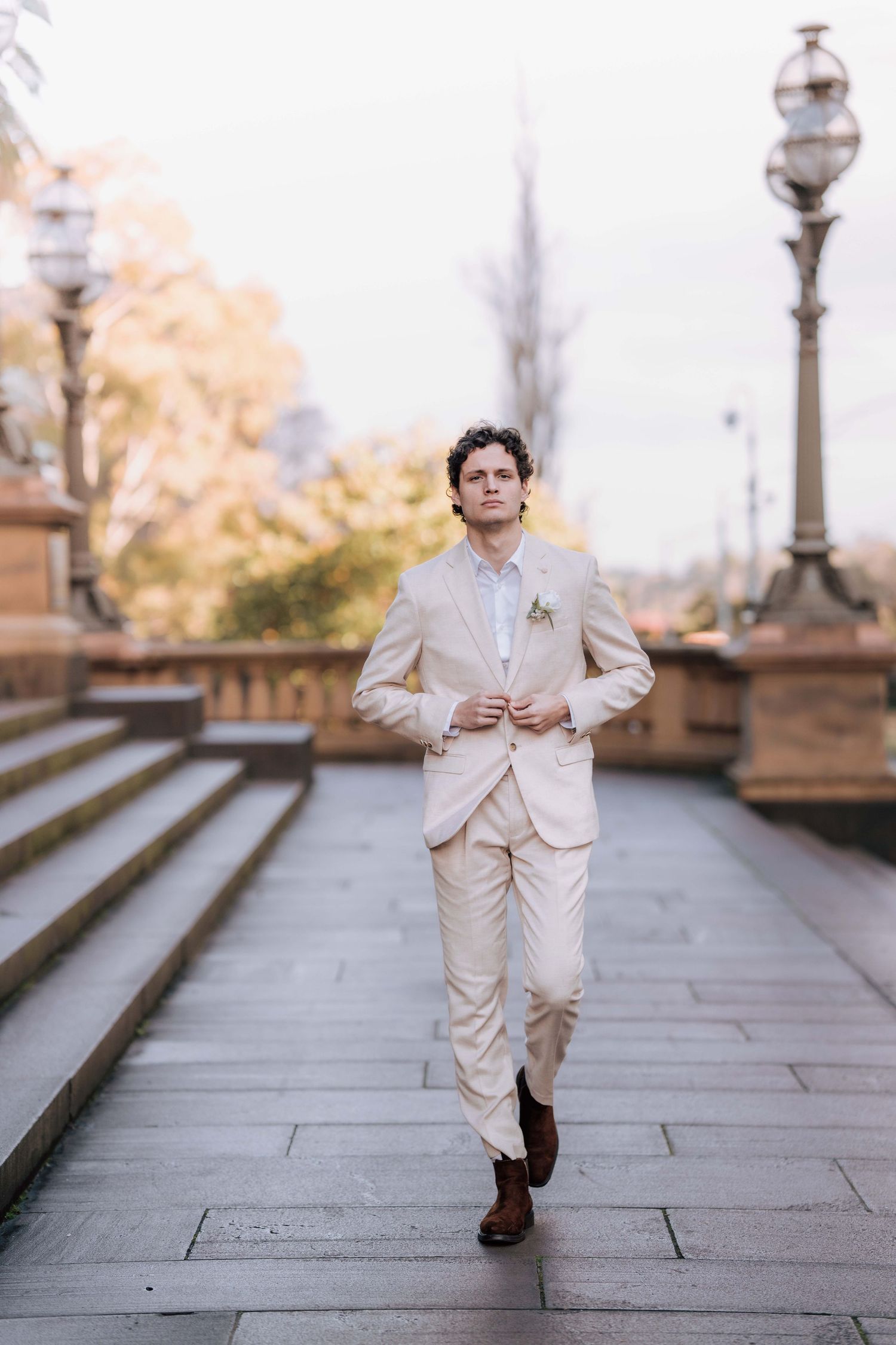 Dapper groom in beige suit with boutonniere walks confidently on stone pathway near steps and vintage lamp posts.