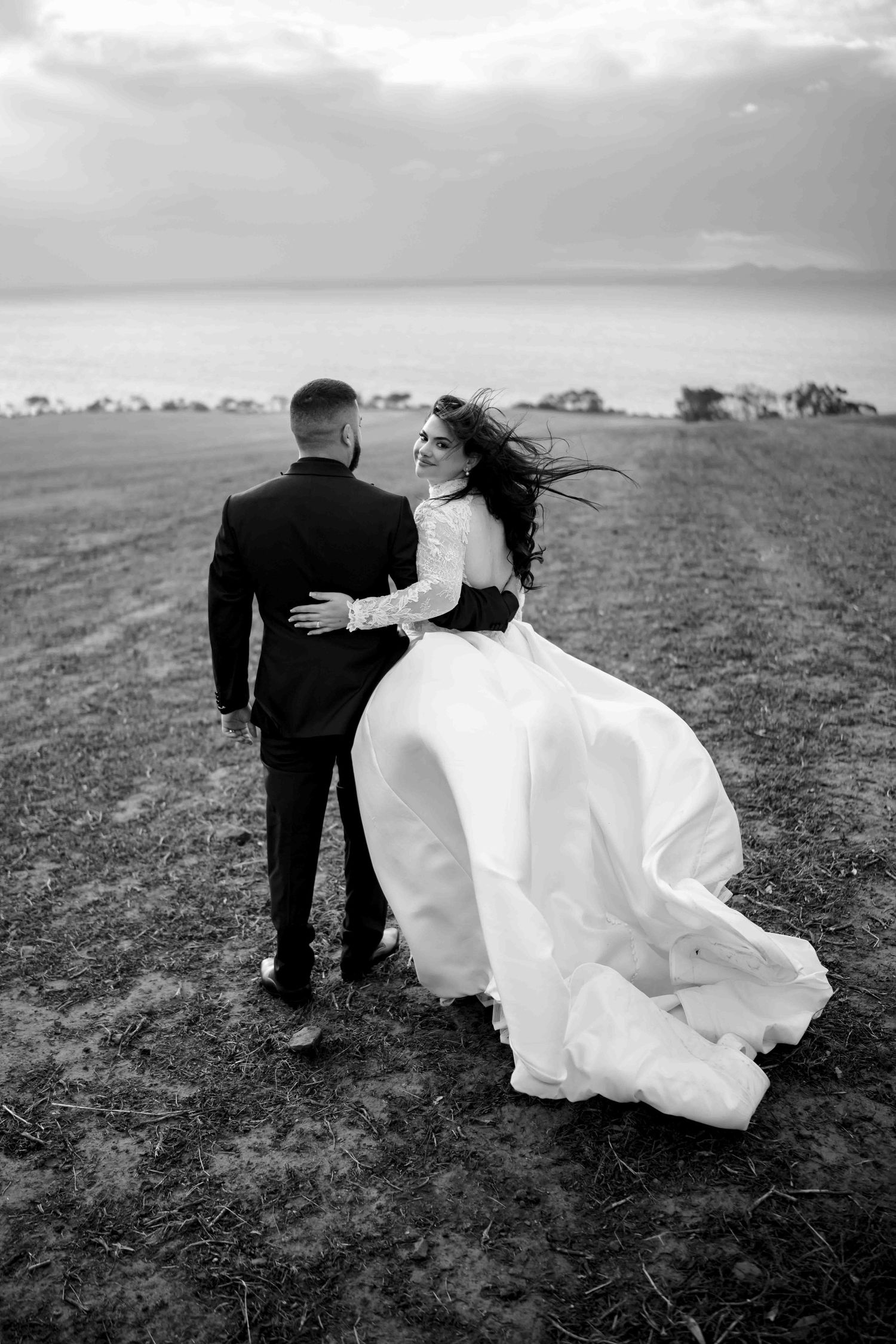 Black and white photo of bride and groom embracing in a field, bride's gown flowing in the wind near the ocean.