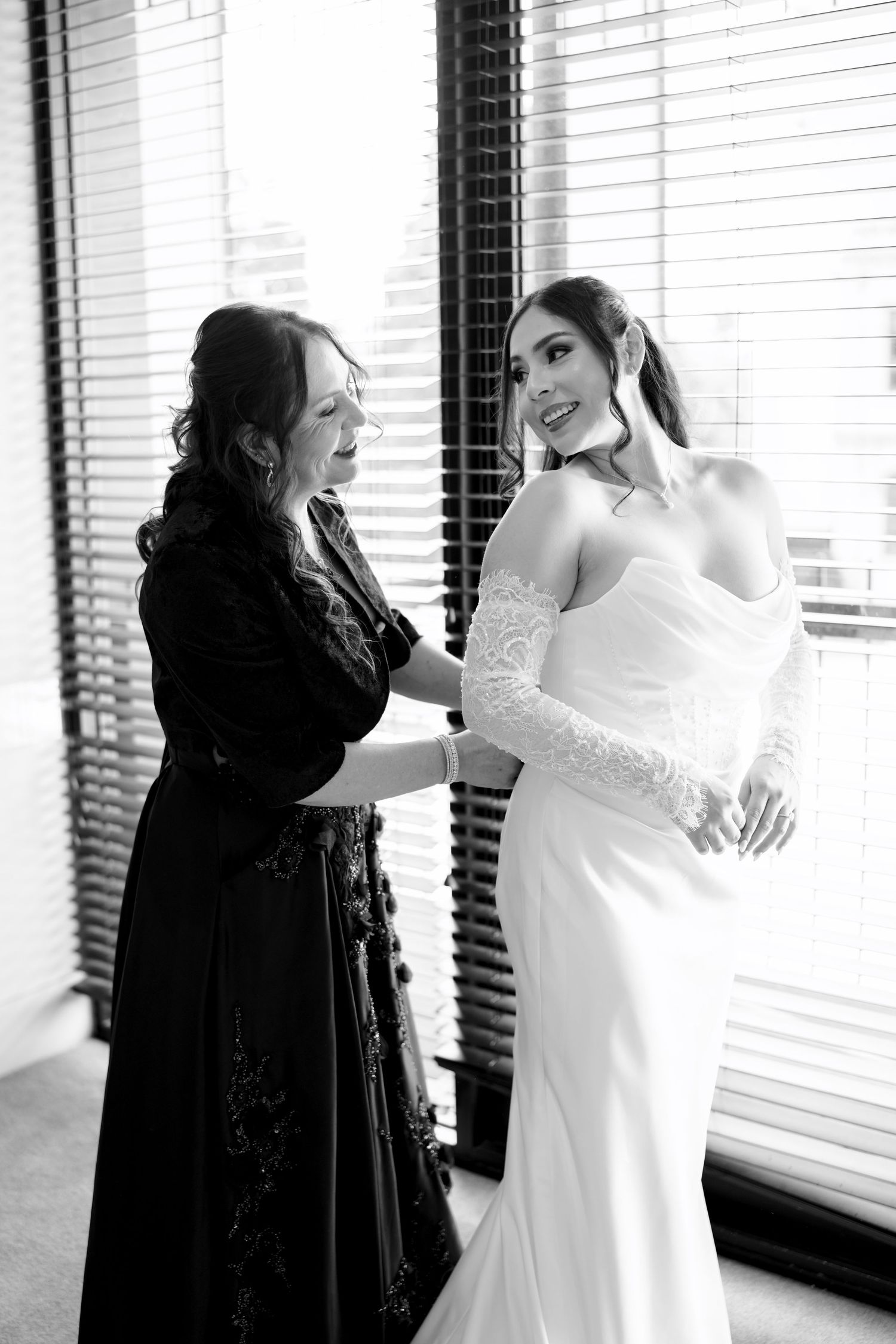 Black and white image of a woman helping a bride in a fitted white lace wedding gown get dressed.
