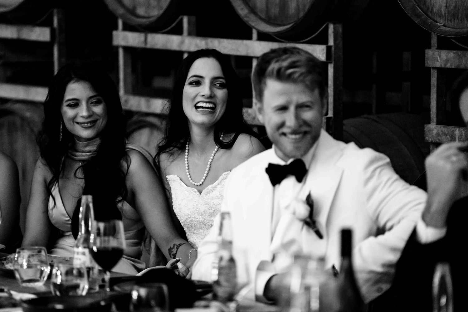 Black and white photo of laughing bride and groom seated at wedding reception table with guests.