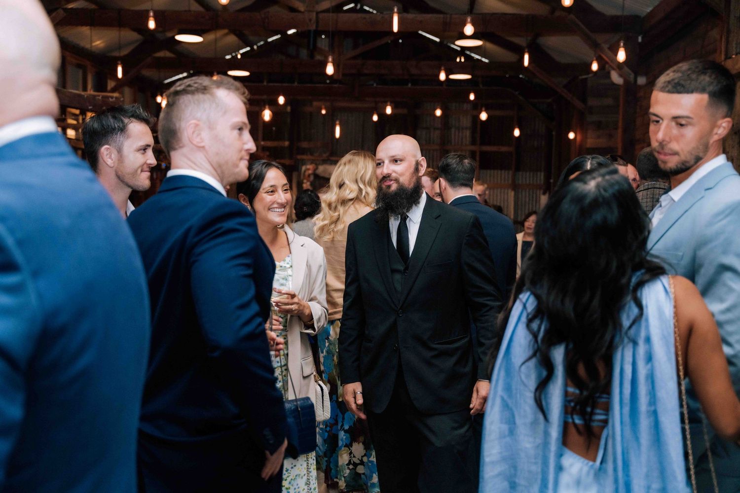 Guests in formal attire mingle and converse inside a warmly lit wedding reception venue.