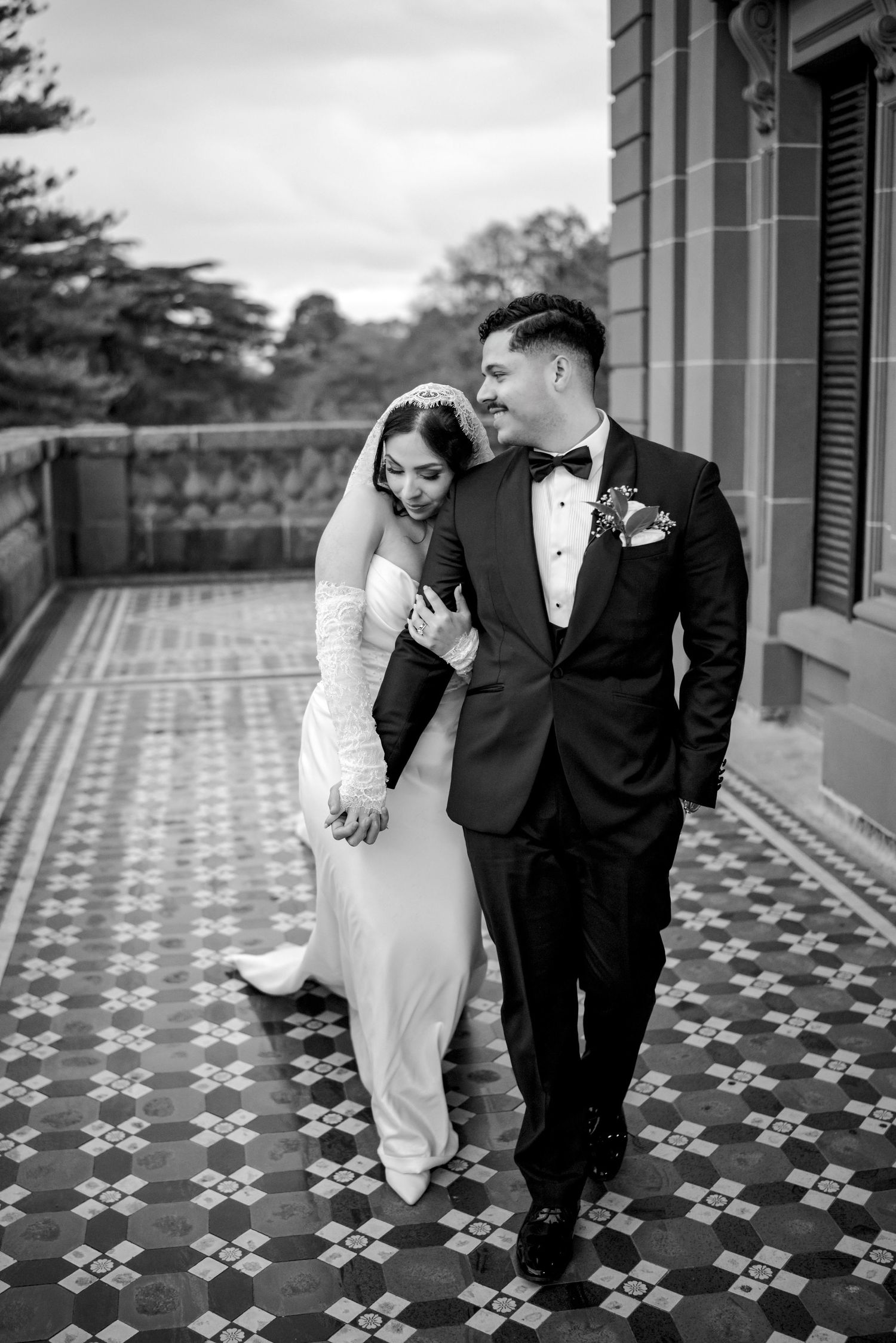 Black and white photo of bride leaning on groom as they walk on a decorative tiled balcony.