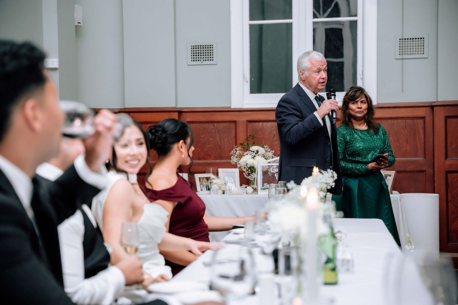 Older gentleman delivers wedding reception toast at microphone while guests seated at formal dinner table listen.