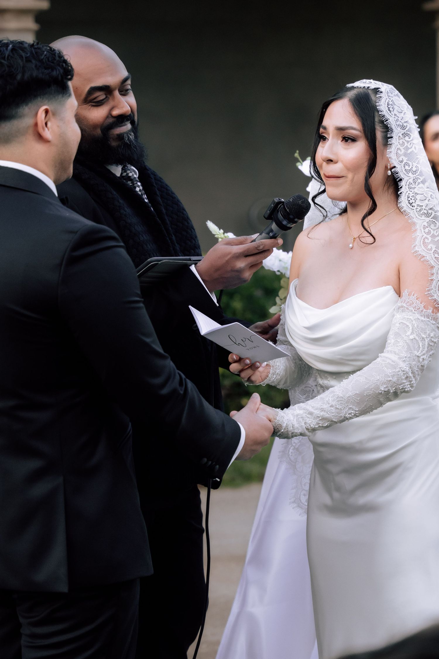 Bride reads wedding vows while holding groom's hands during outdoor ceremony with officiant.