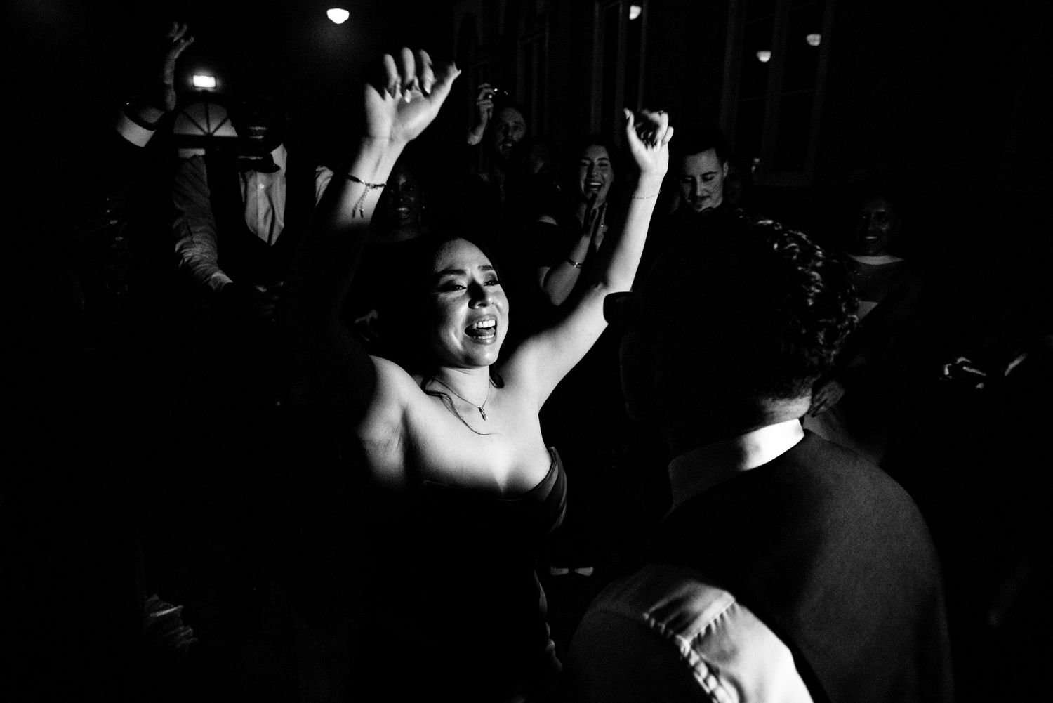 Black and white photo of a woman in a strapless dress dancing joyfully at a wedding reception.