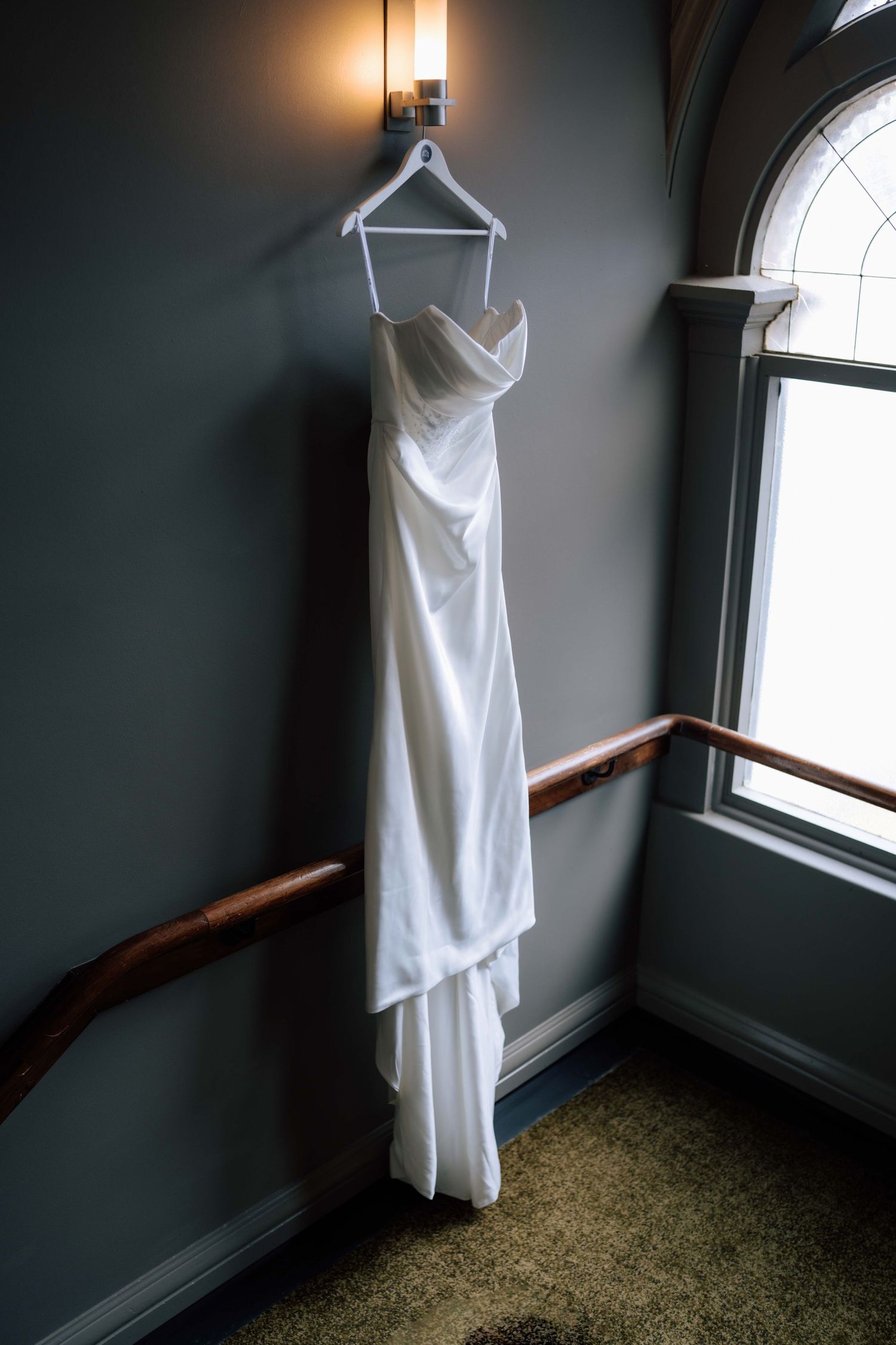 White strapless wedding dress hanging on a hanger against a grey wall near a window.