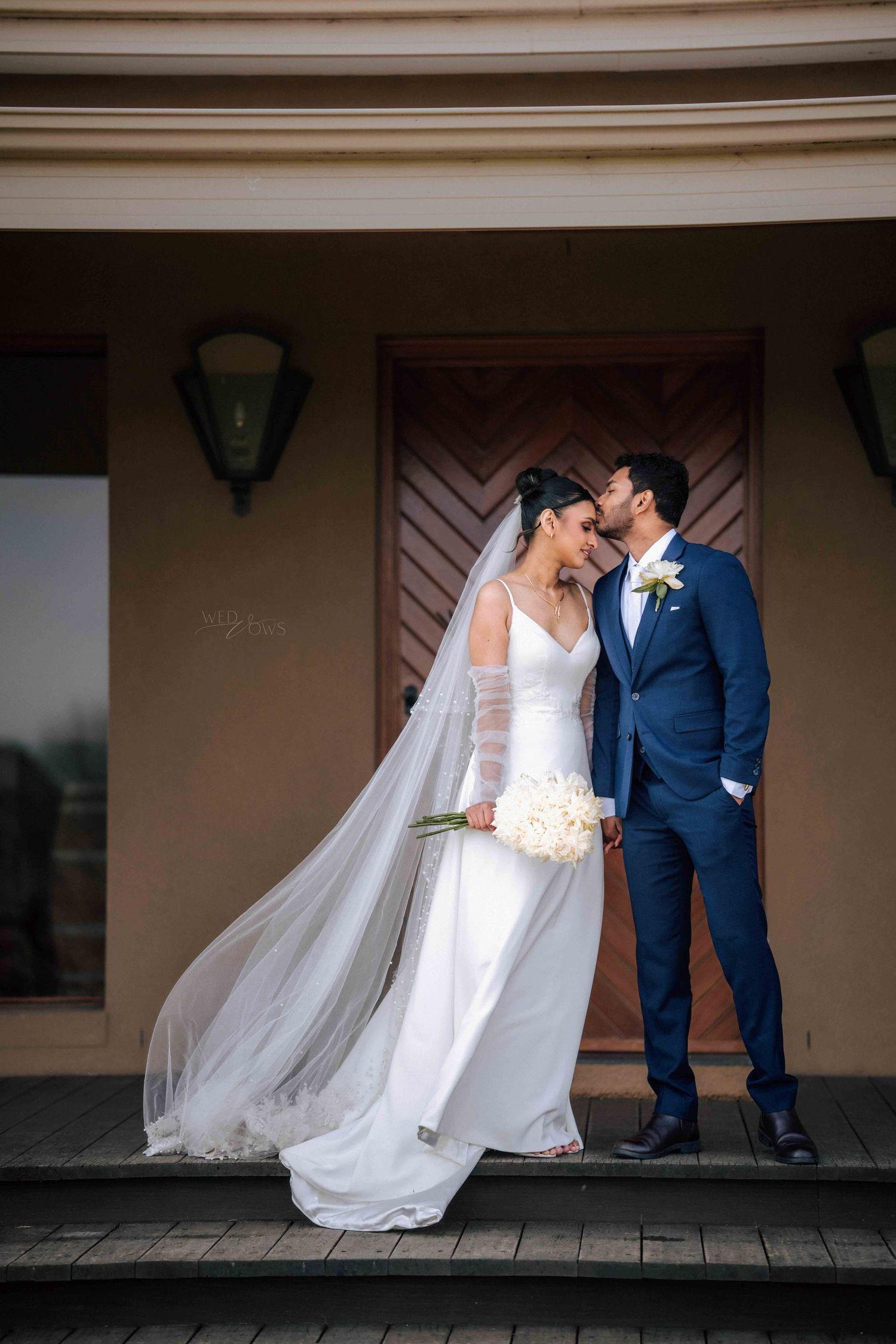 Groom in navy suit kissing bride on cheek outside venue, her long veil flowing in the breeze.