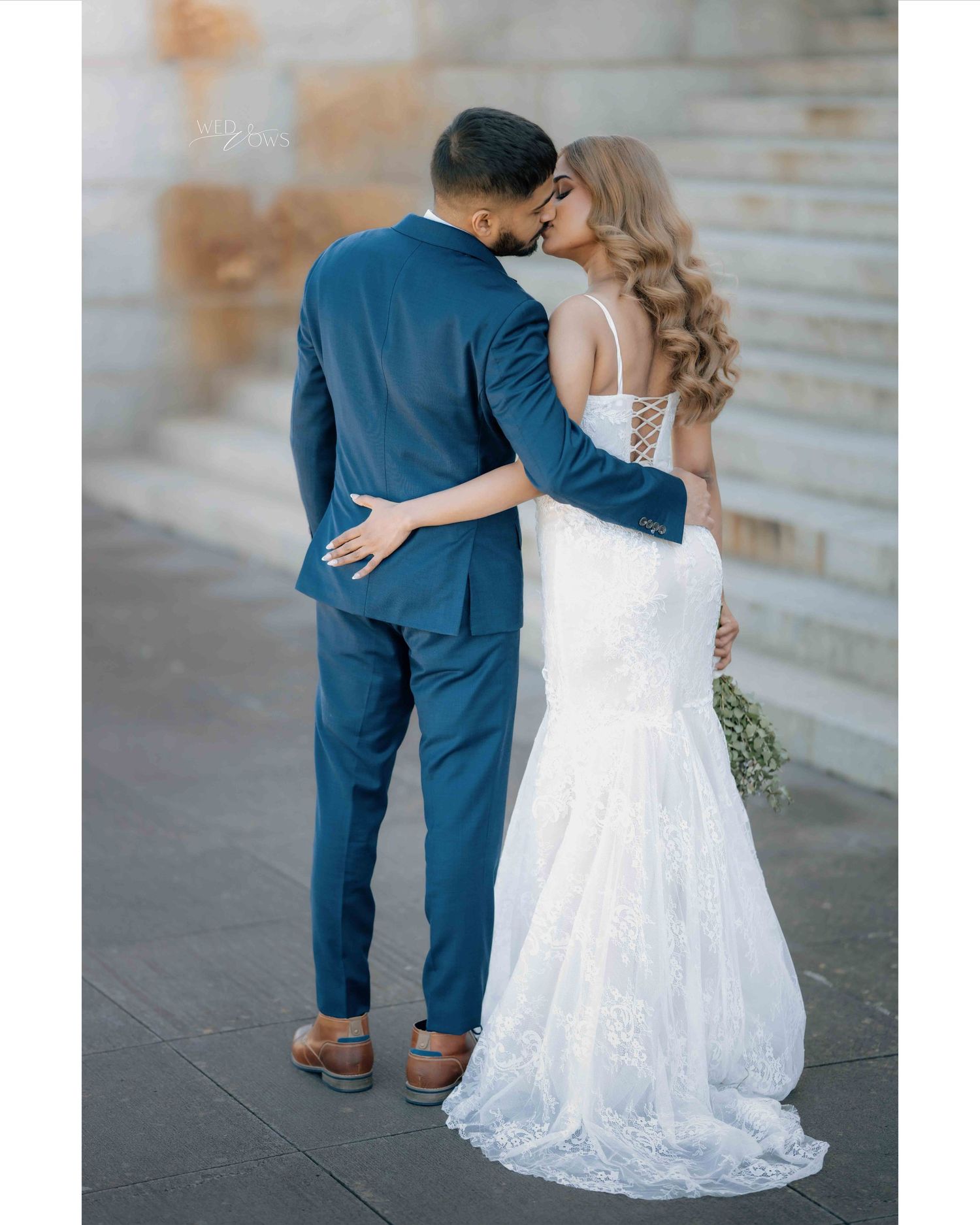 Bride and groom sharing a kiss, showcasing lace back wedding gown and navy blue suit.