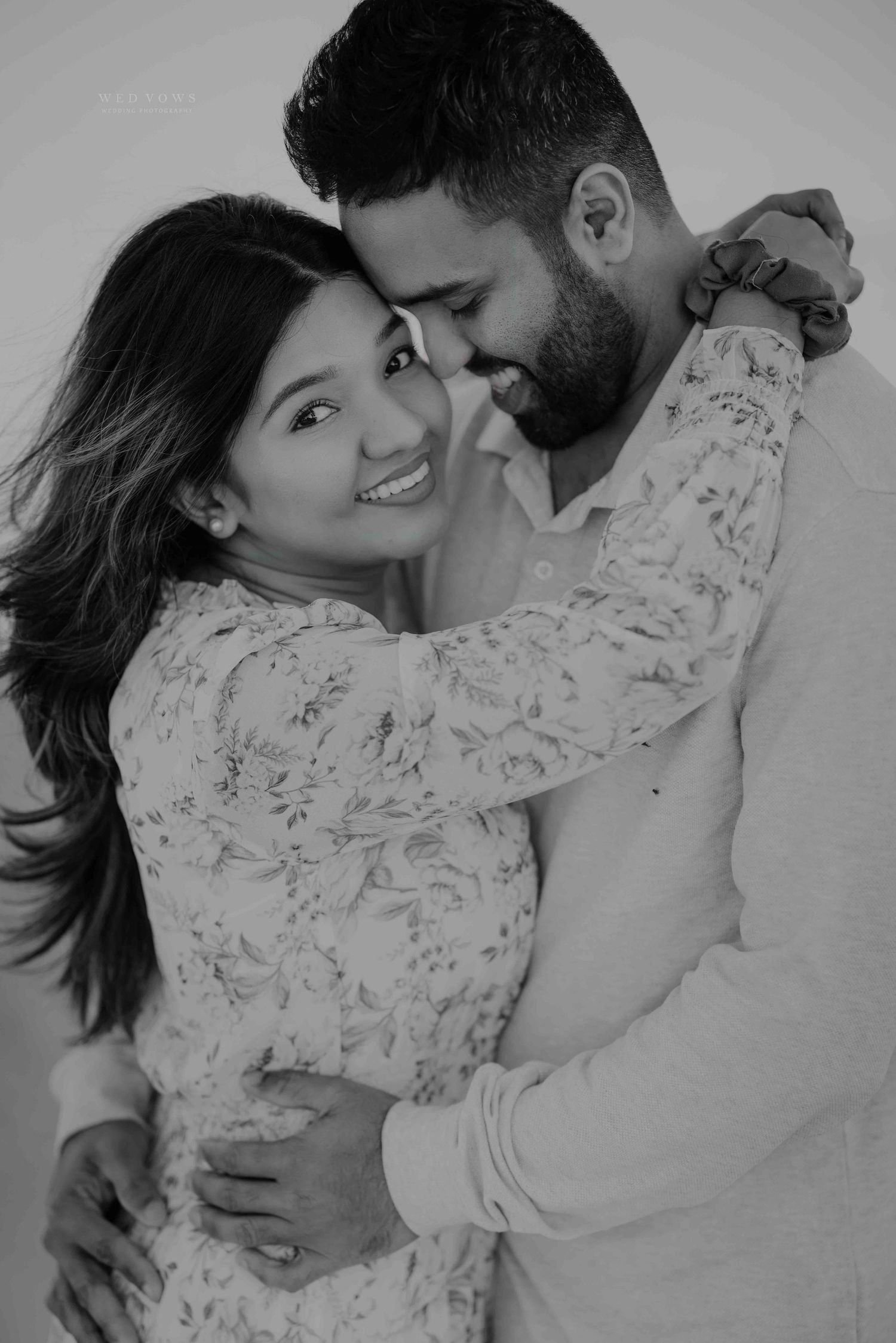 Black and white portrait of a couple embracing tenderly, woman smiling as man nuzzles her forehead.