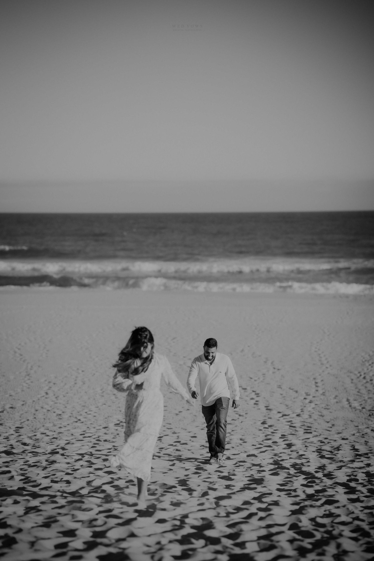 Black and white photo of couple holding hands walking on sandy beach with ocean waves in background.