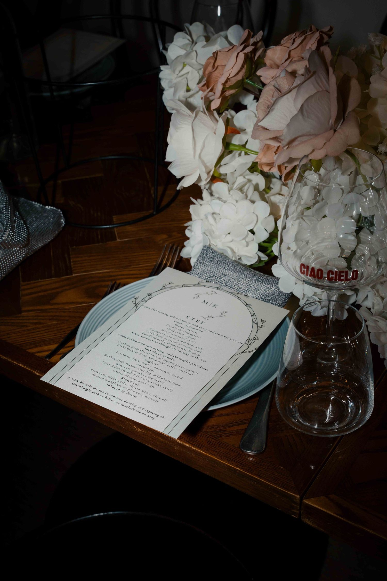 Elegant wedding table setting with floral centerpiece, menu card on blue plate, and Ciao Cielo water bottle.