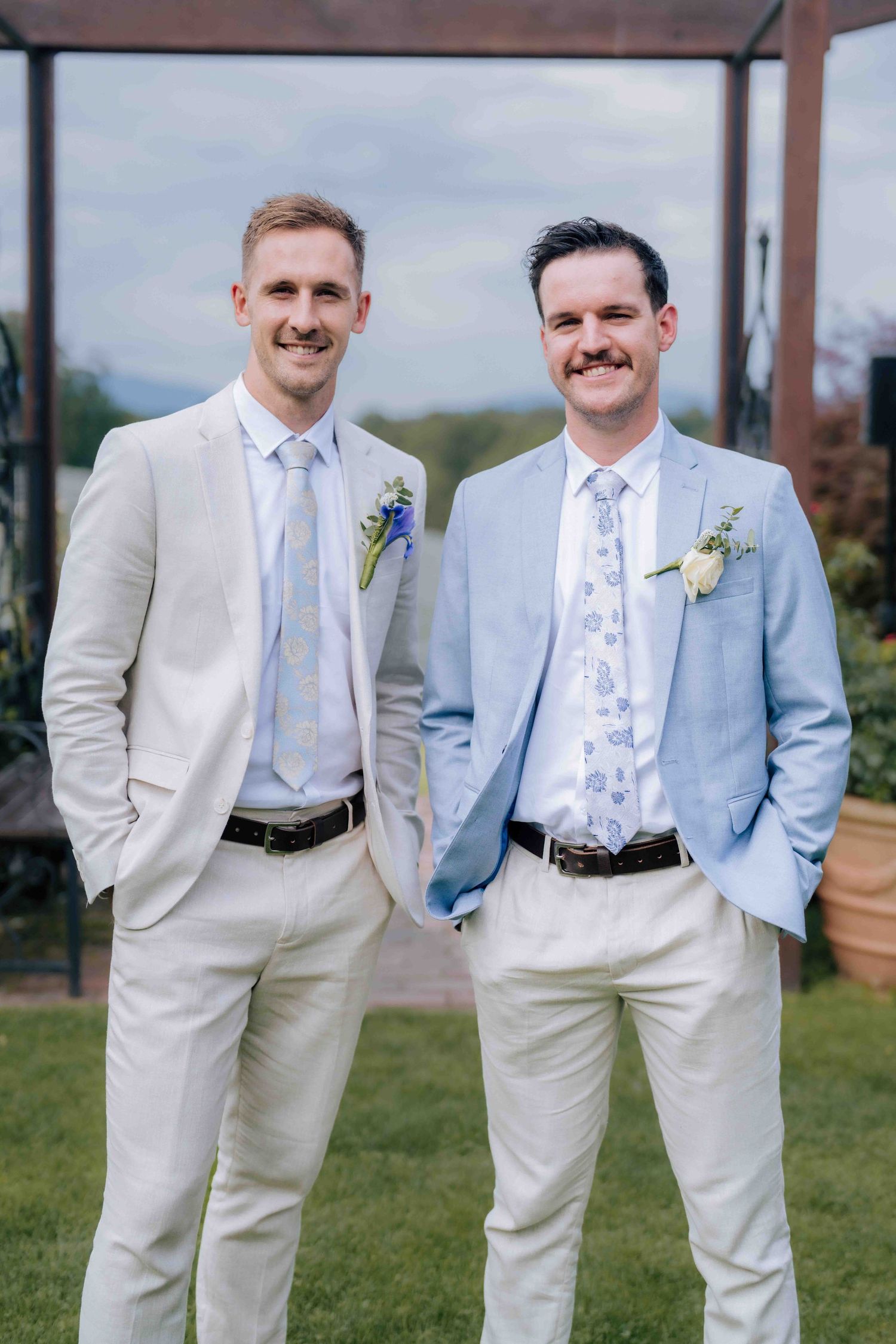 Two groomsmen in light suits with boutonnieres smile outdoors at a wedding ceremony.