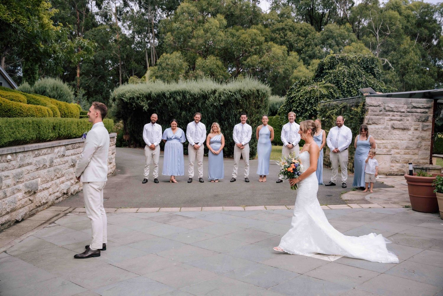 Bride and groom share first look moment on stone patio with wedding party watching in background.