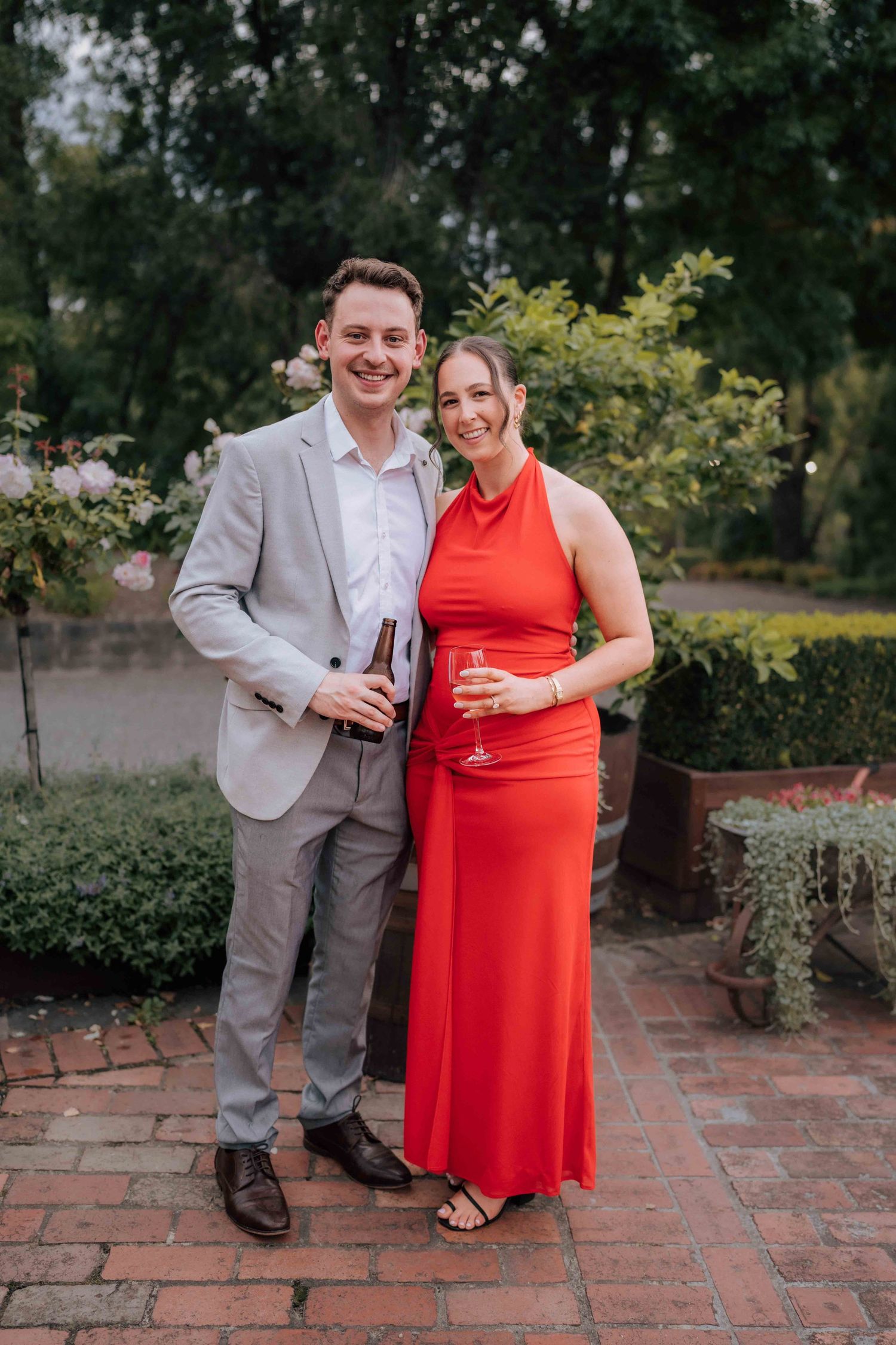 Couple in formal attire holding champagne glasses pose in an outdoor garden setting.