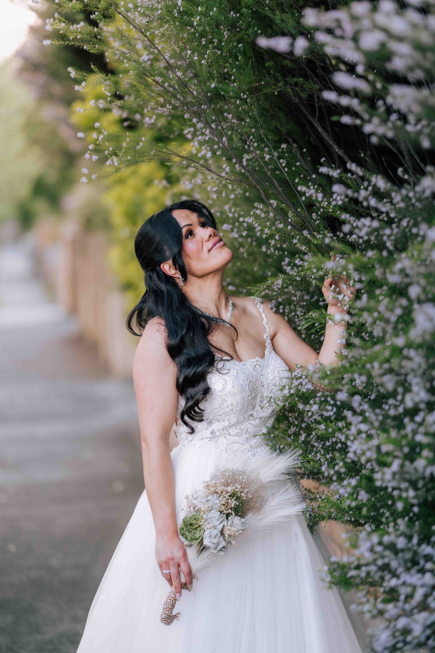 Bride in white ballgown gazing upward while standing among flowering garden bushes.