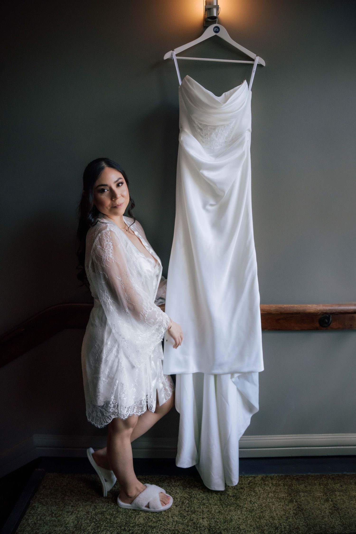 Bride in lace robe poses beside her elegant strapless wedding gown hanging in a dimly lit room.