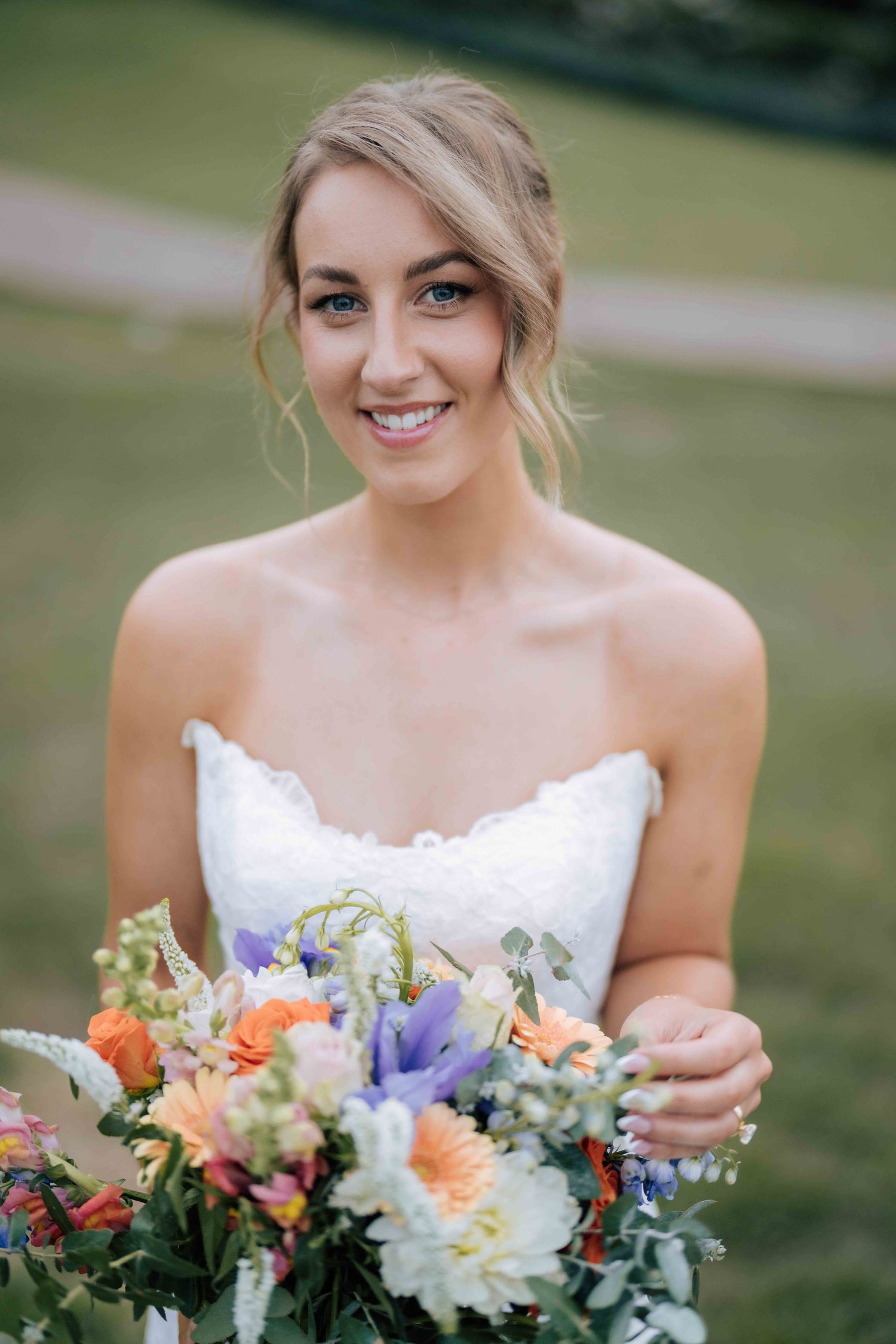 Beautiful blonde bride in strapless white lace dress smiles warmly while holding a vibrant colorful floral bouquet.