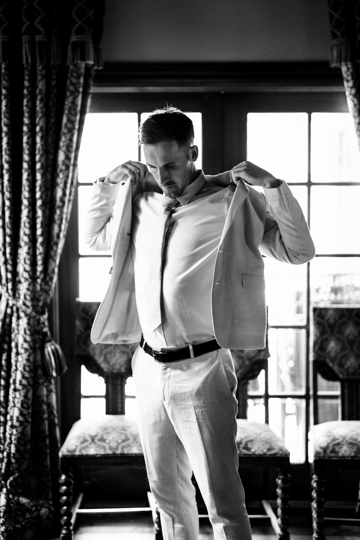 Dapper groom in white suit adjusting jacket collar in black and white photo by window.