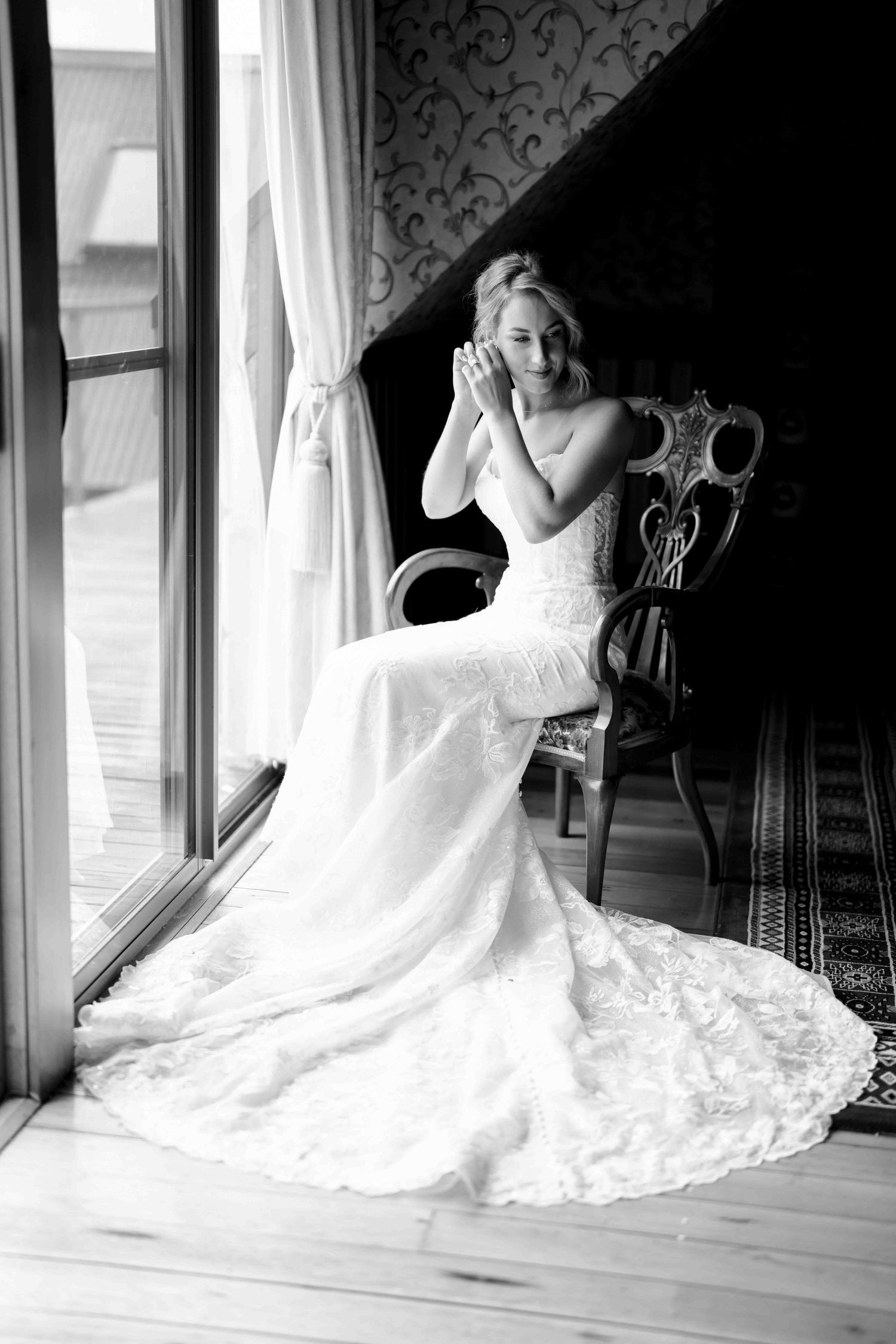 Black and white portrait of emotional bride in fitted lace gown seated by window with hands clasped.