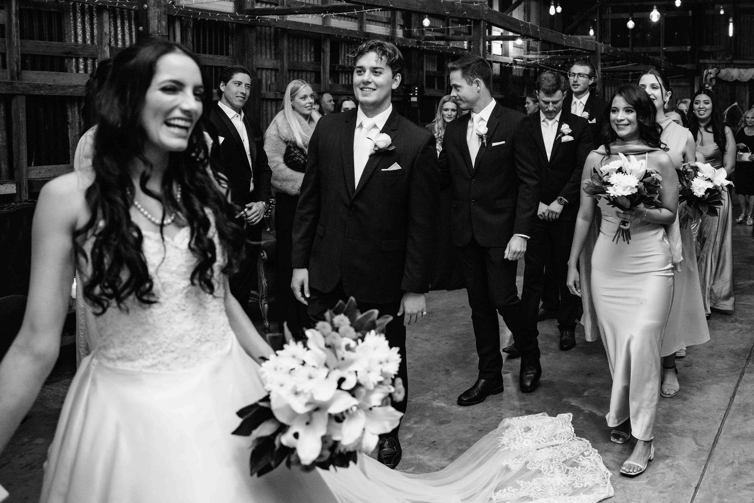 Black and white photo of smiling bride with bouquet and wedding party walking through a rustic barn venue.