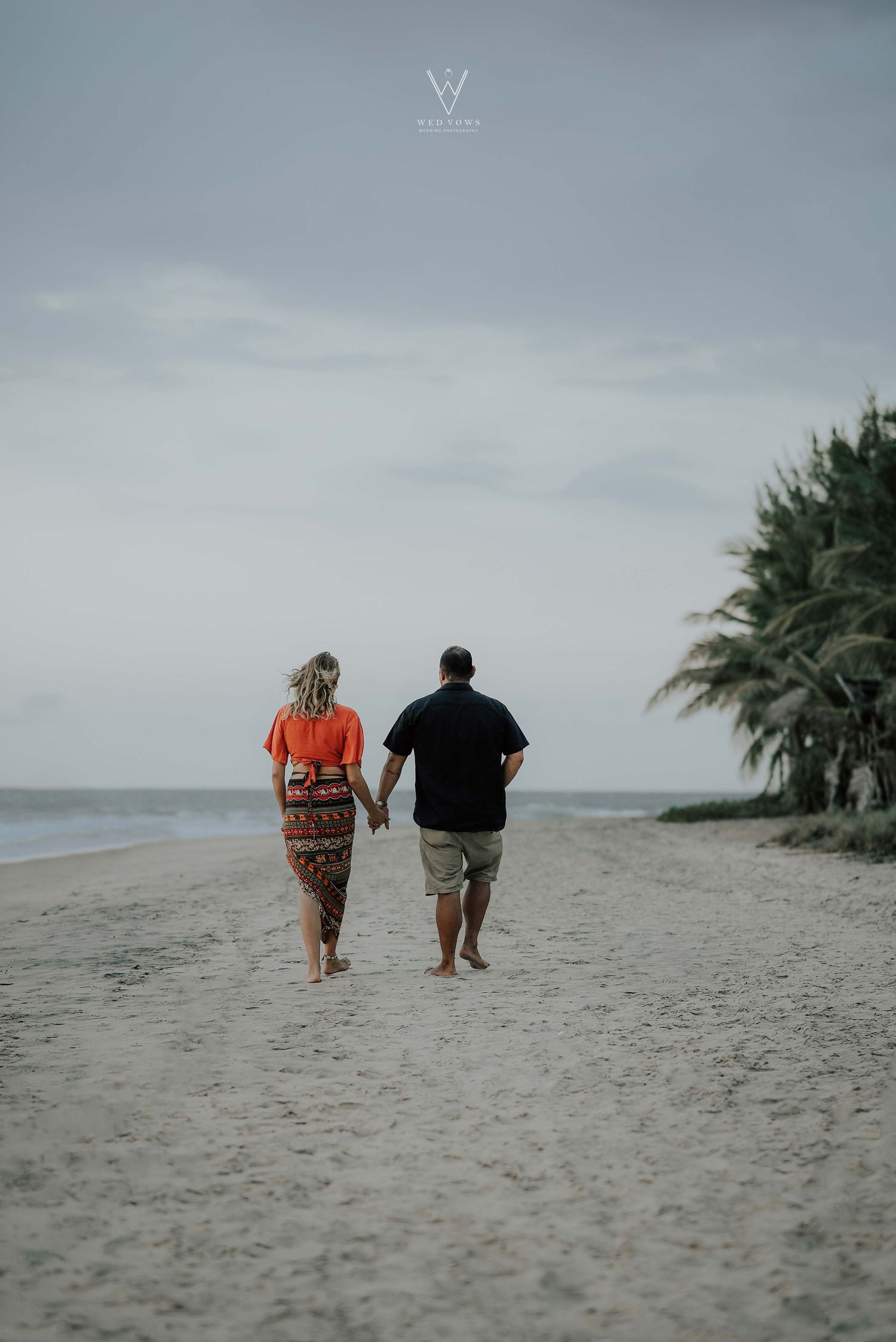 Couple walking hand-in-hand along a tropical beach lined with palm trees under a cloudy sky.