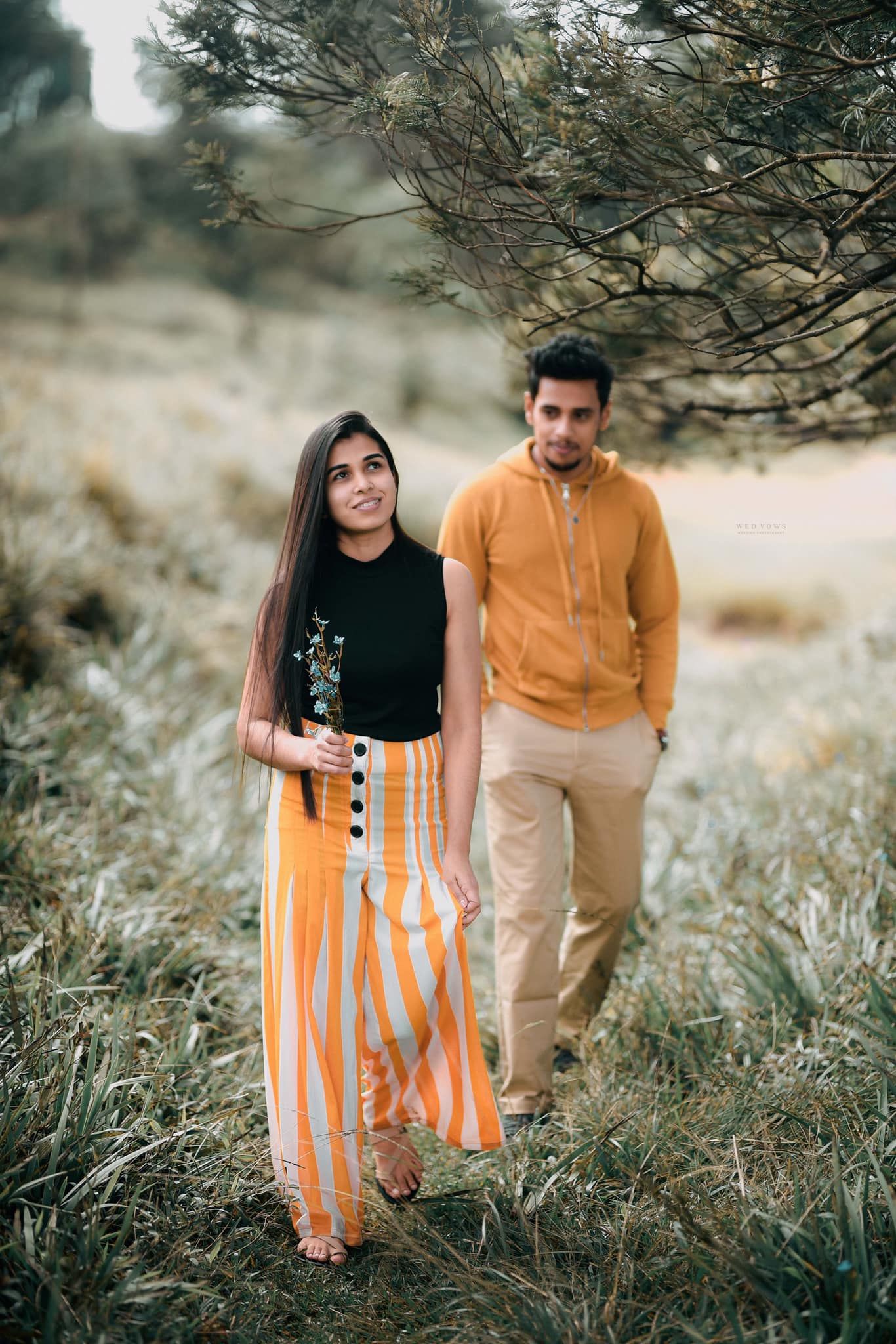 Young couple posing in grassy field, woman in striped orange pants and black top.