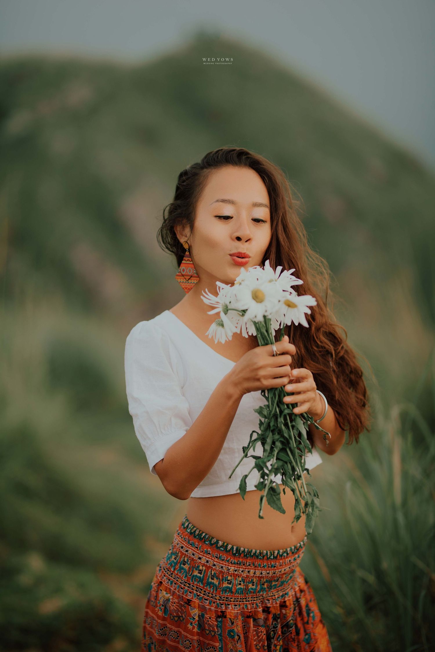 Young woman holding white daisy bouquet with green hills in background.