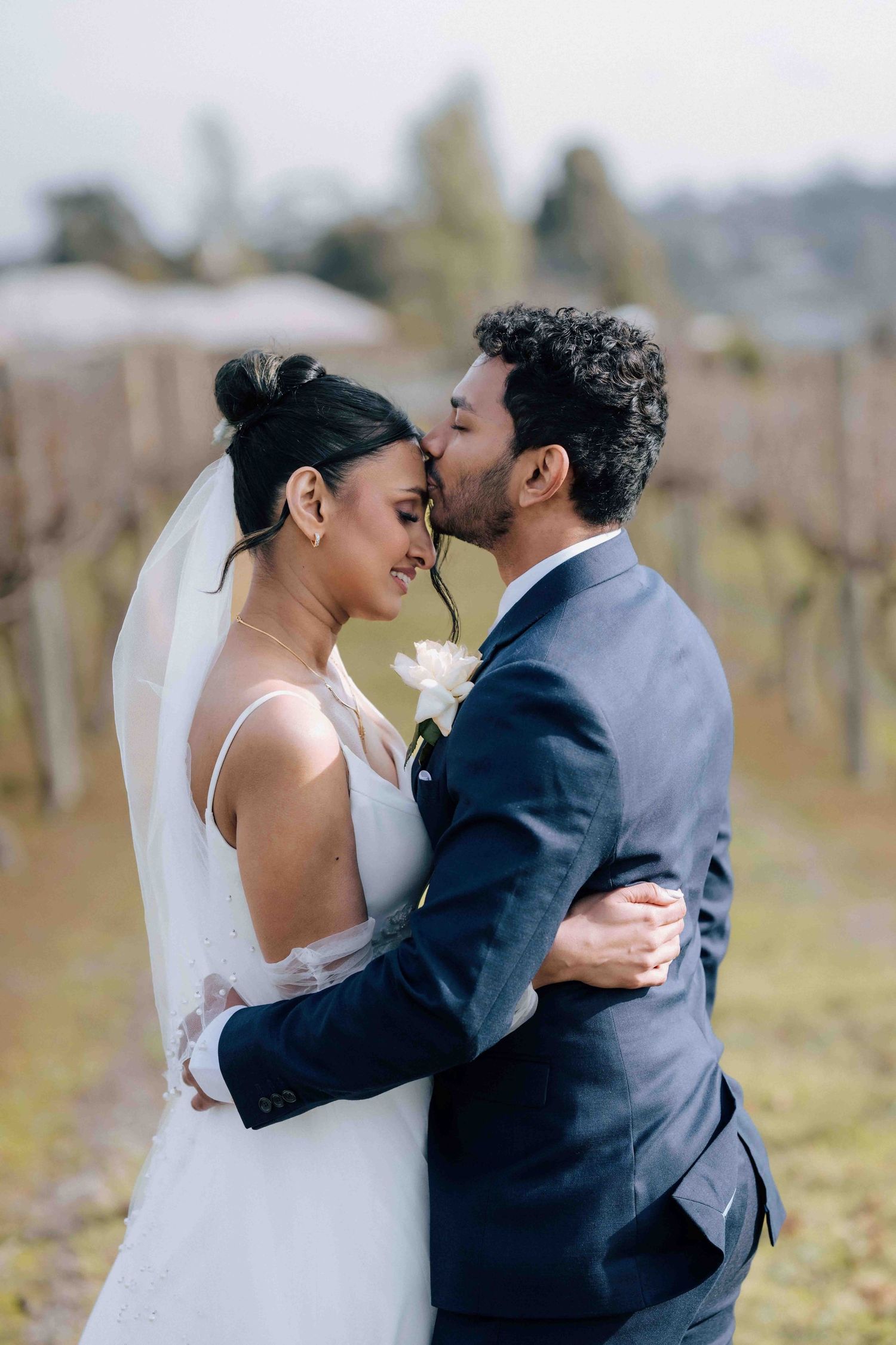 Groom kisses bride on the forehead as they embrace in a vineyard on their wedding day.