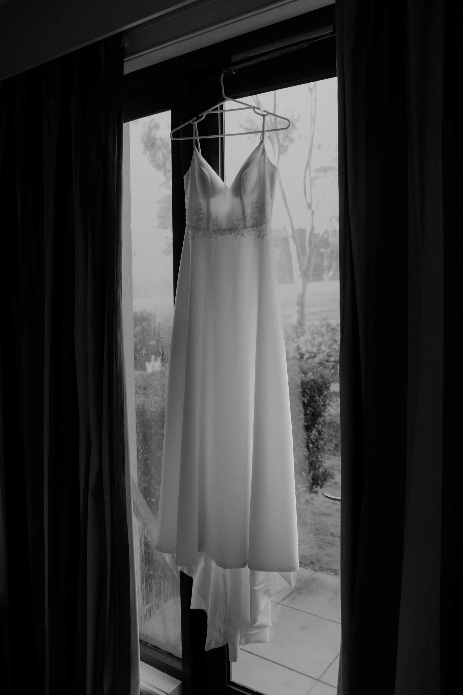 Black and white photo of elegant spaghetti strap wedding gown hanging in window with natural light.
