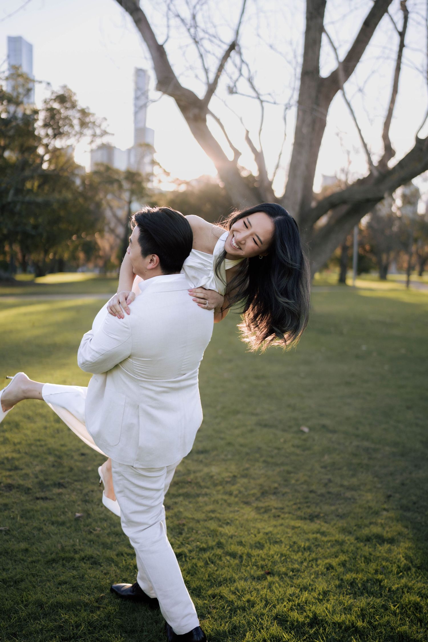 Groom in white suit lifts and spins laughing bride in white dress on green park lawn at golden hour.