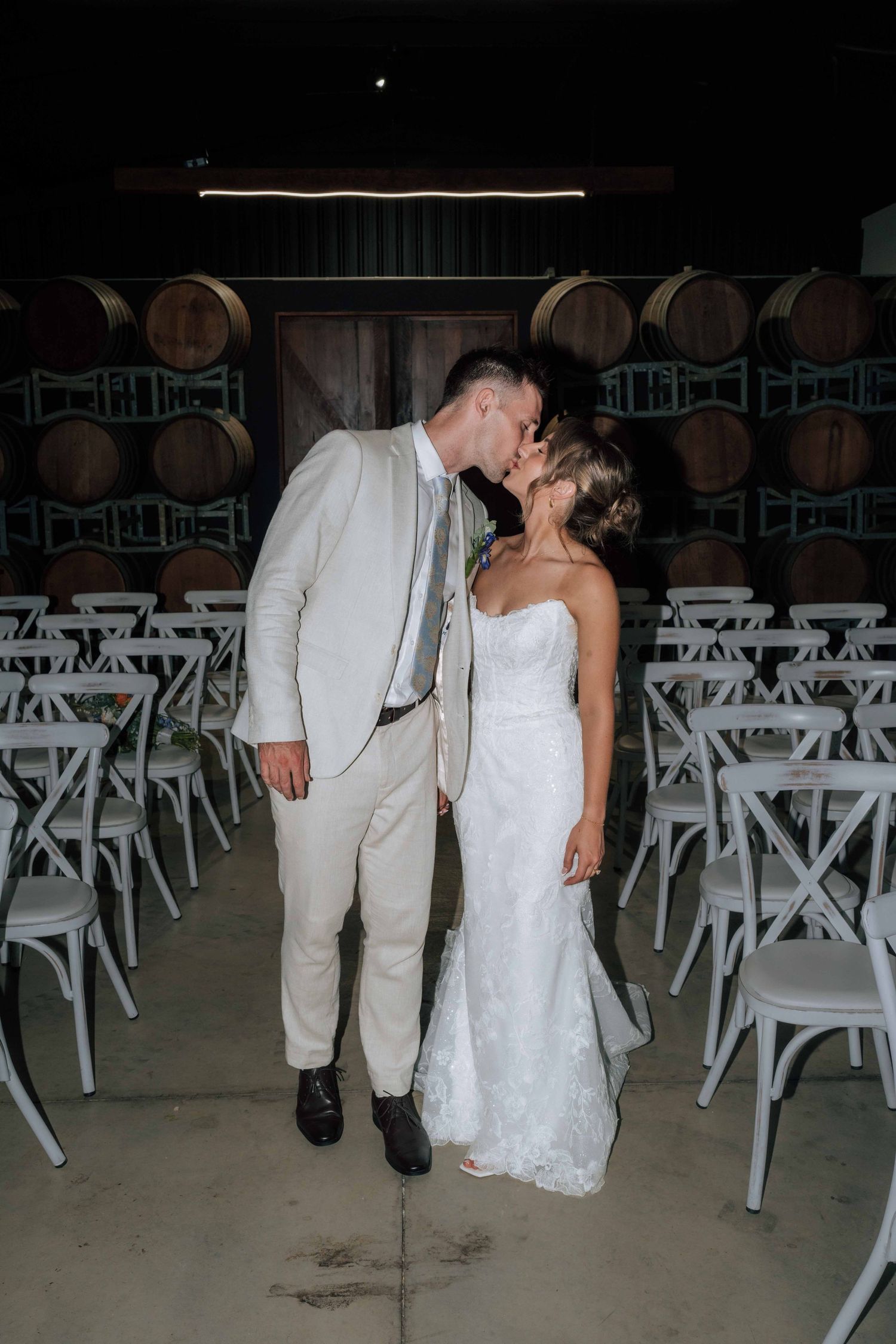 Bride and groom share tender kiss surrounded by wine barrels and white cross-back chairs in winery venue.