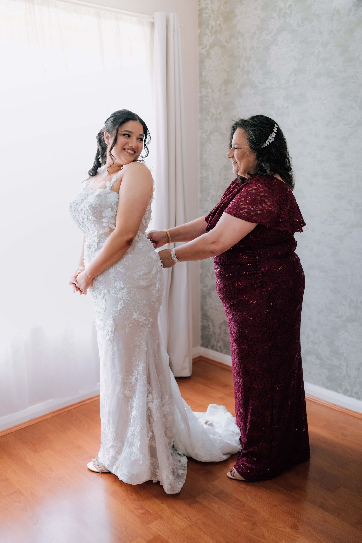 Mother in burgundy lace dress helps bride in fitted lace wedding gown get dressed by bright window.