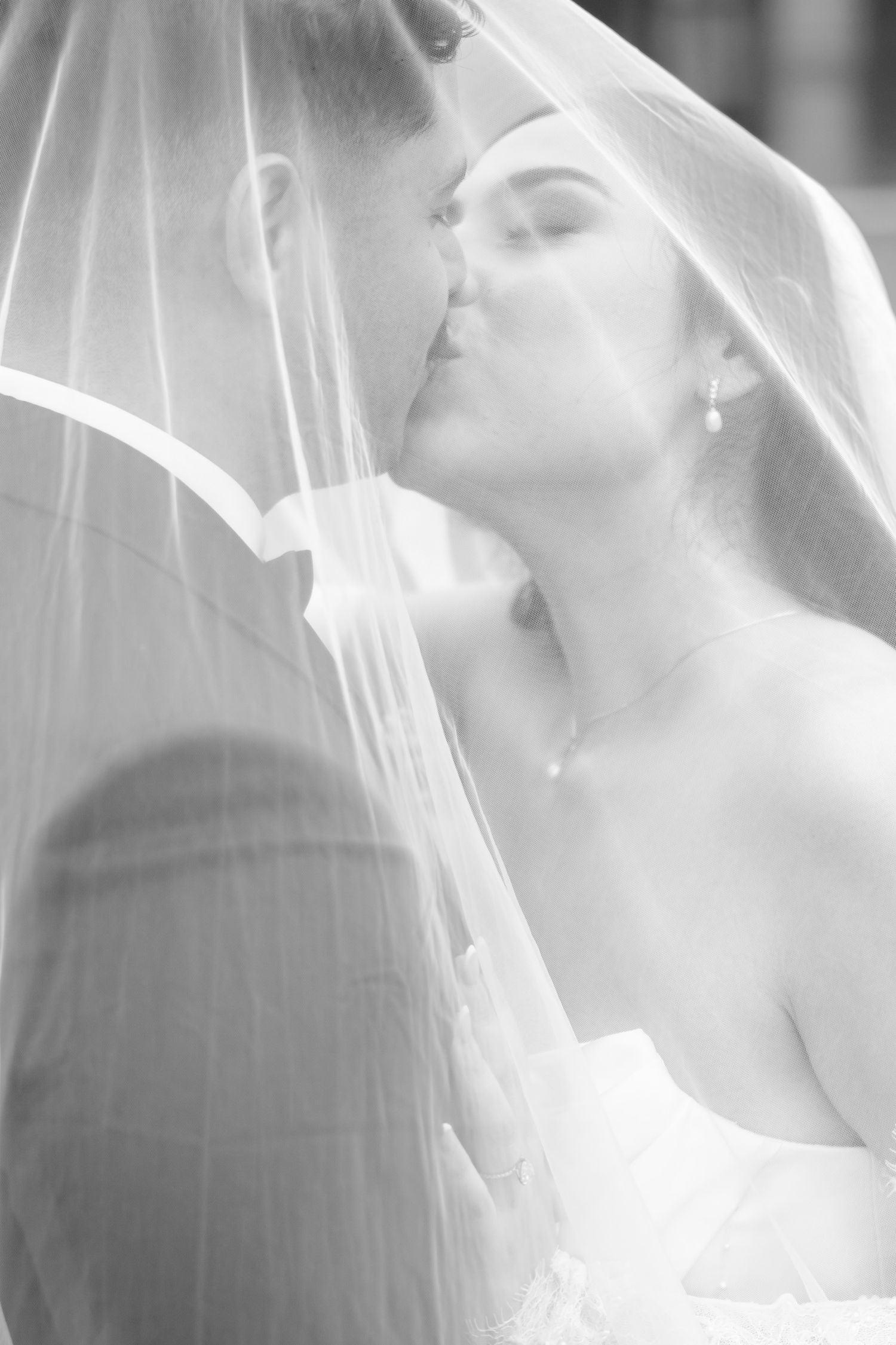 Black and white romantic photo of bride and groom kissing under a flowing wedding veil.